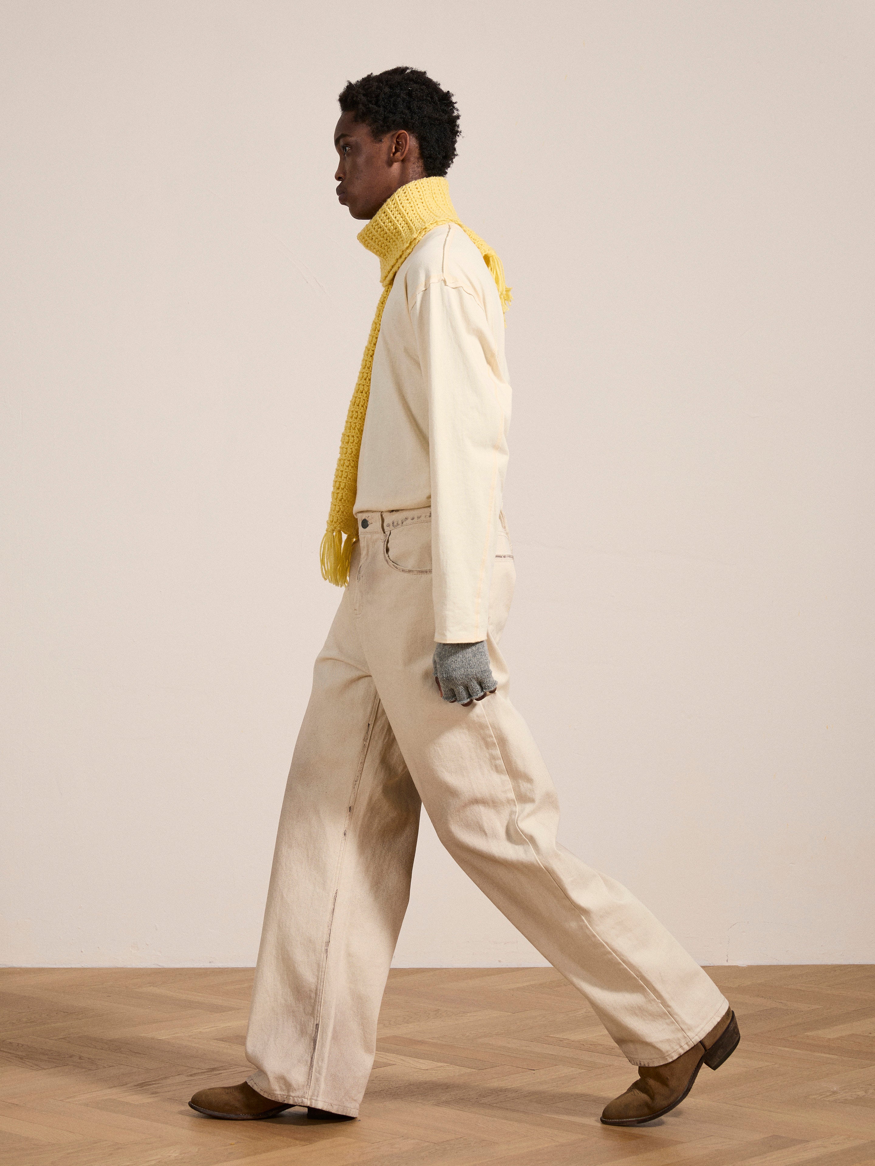 A person wearing a light beige long-sleeve shirt, Lacy Baggy Jeans by FOUND in dirty white wash, brown boots, grey gloves, and a yellow scarf walks on a wooden floor against a plain off-white background.