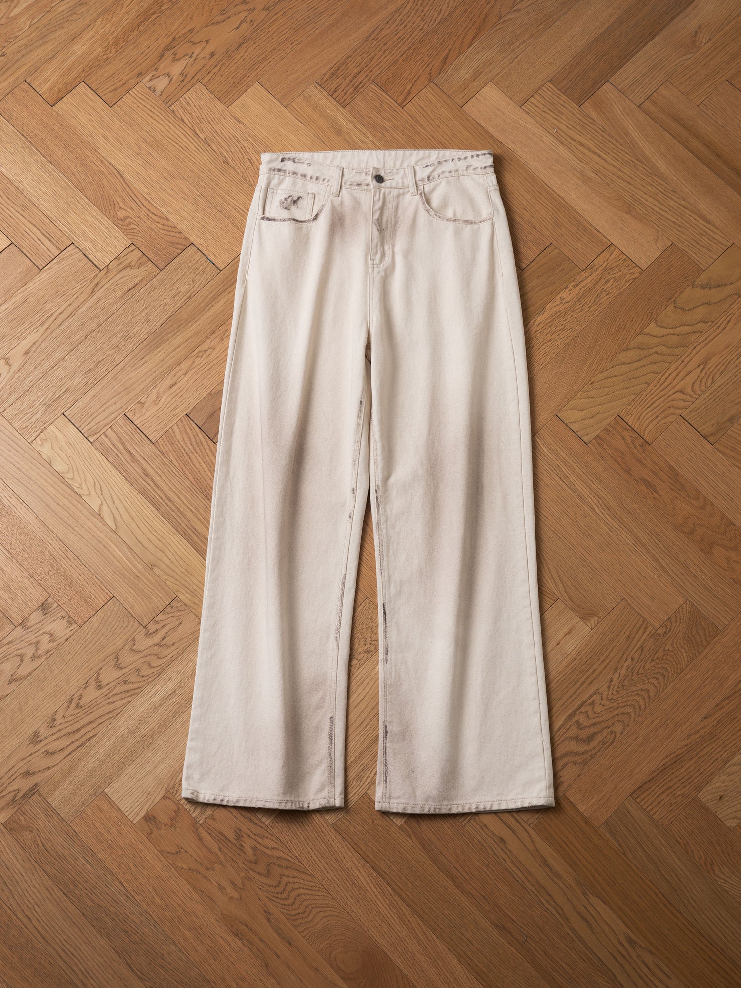 A pair of Lacy Baggy Jeans by FOUND, featuring a beige color with wide-leg cut and light distressing, along with front and back pockets and a single button closure, laid flat on a wooden herringbone patterned floor.