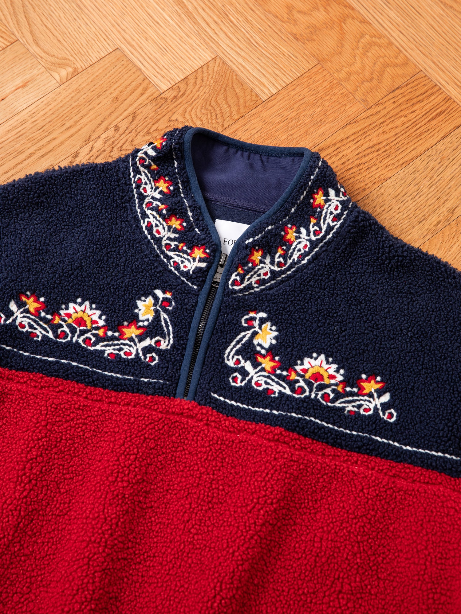 A close-up of the FOUND Pahari Floral Embroidered Sherpa Pullover, featuring elegant navy blue and red fleece adorned with intricate floral embroidery near the collar and chest area, laid out on a wooden herringbone floor.