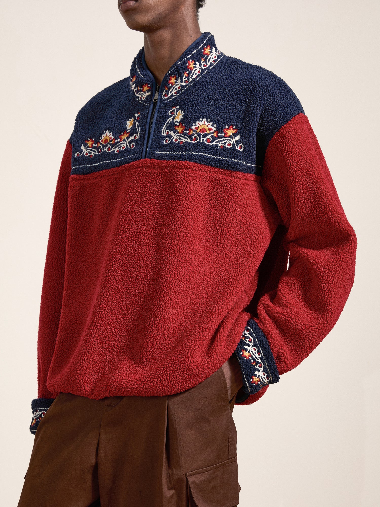 Individual dressed in a red and navy blue FOUND Pahari Floral Embroidered Sherpa Pullover, paired with brown pants.