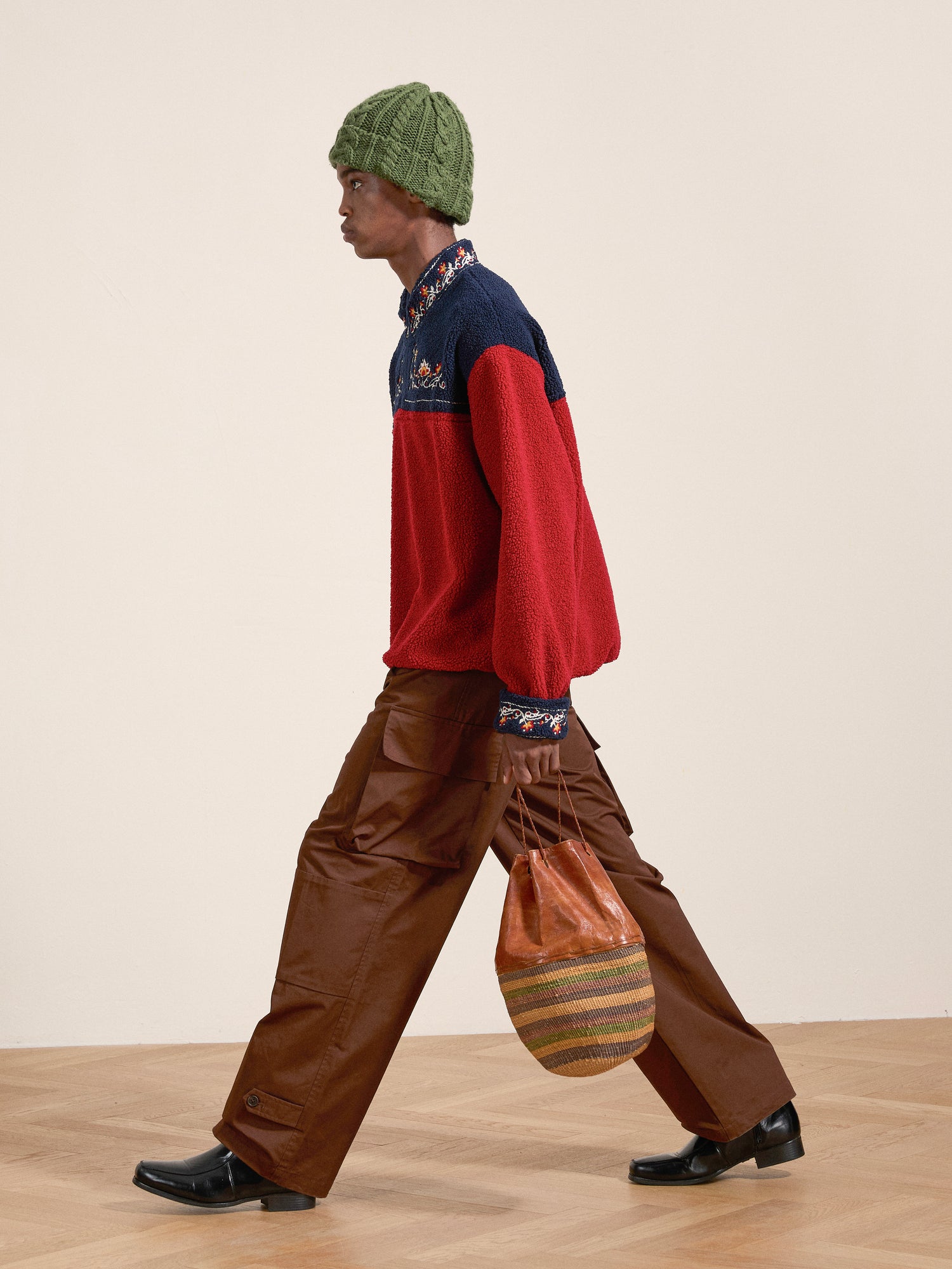 A person walks indoors carrying a round, woven bag. They wear a green knit hat, the Pahari Floral Embroidered Sherpa Pullover by FOUND in red and navy, brown pants with pockets, and black shoes.