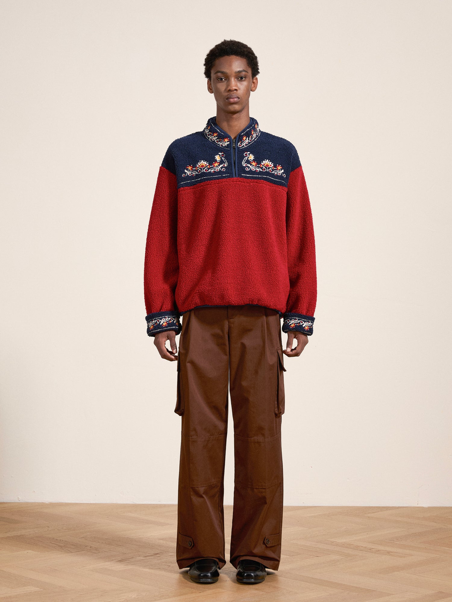 A person stands on a wooden floor, donning FOUND's red and navy Pahari Floral Embroidered Sherpa Pullover, brown cargo pants, and black shoes, with a plain beige background behind them.