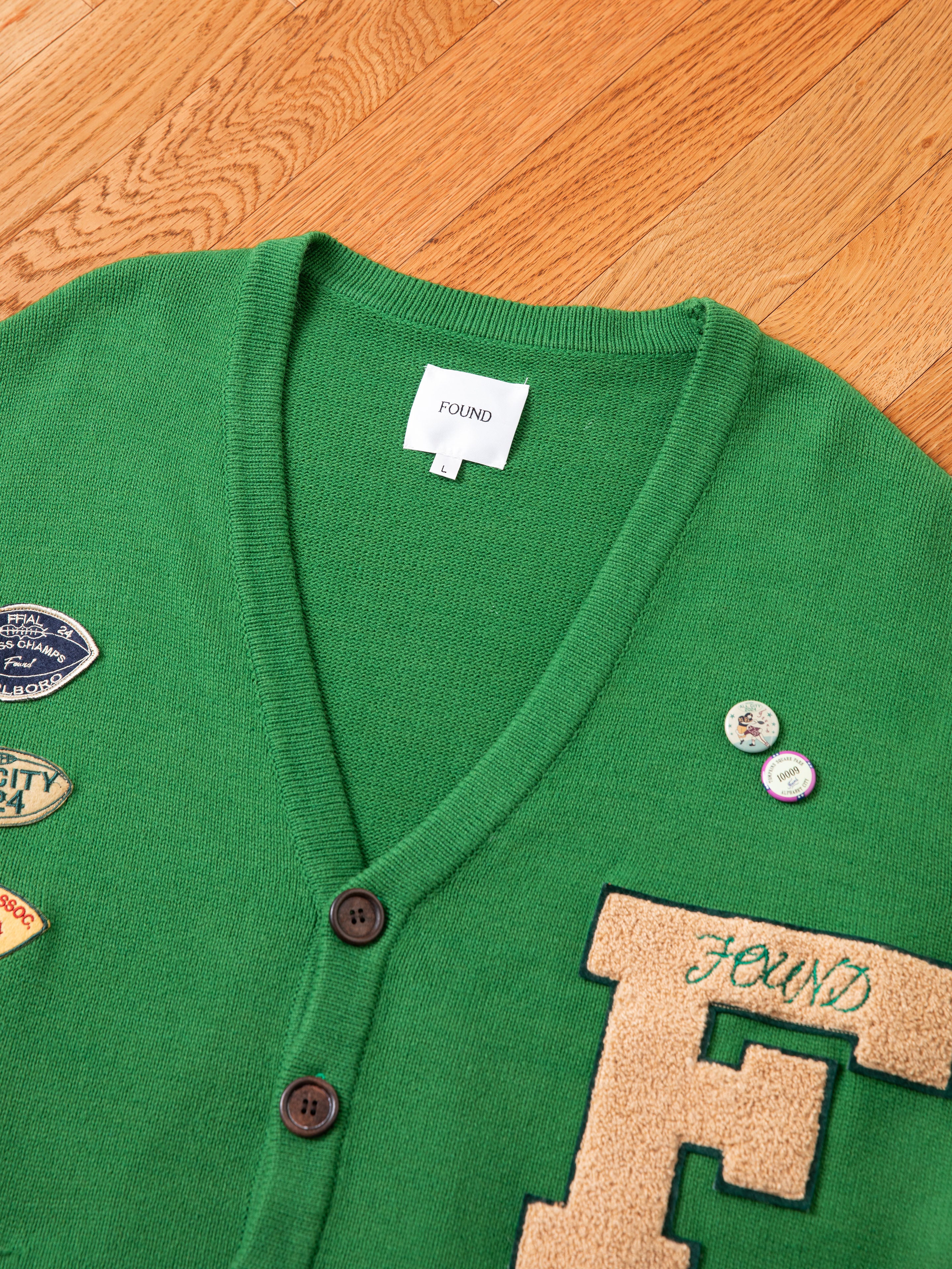 The Pineneedle Varsity Knit Cardigan from FOUND, made of 100% cotton knit, lays on a wooden floor with its green hue and two buttons. Various patches enhance its vintage style, including one large patch featuring the letter "F" and an internal label that reads "FOUND".