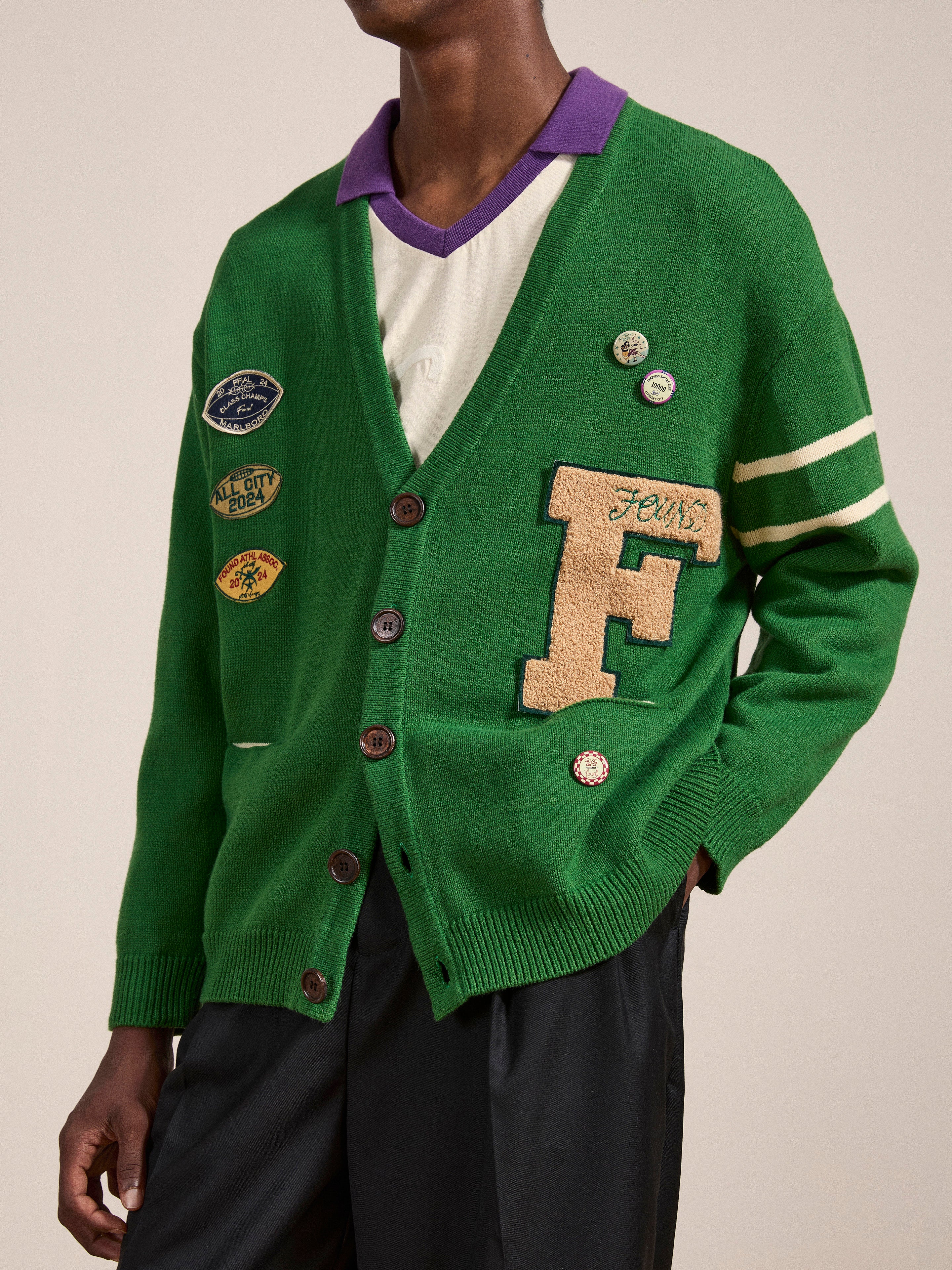 A person is wearing a green Pineneedle Varsity Knit Cardigan by FOUND, which features various patches, a large "F" letter, and buttons. The vintage-styled cardigan is buttoned up over a white shirt with a purple collar. The person stands against a plain background.