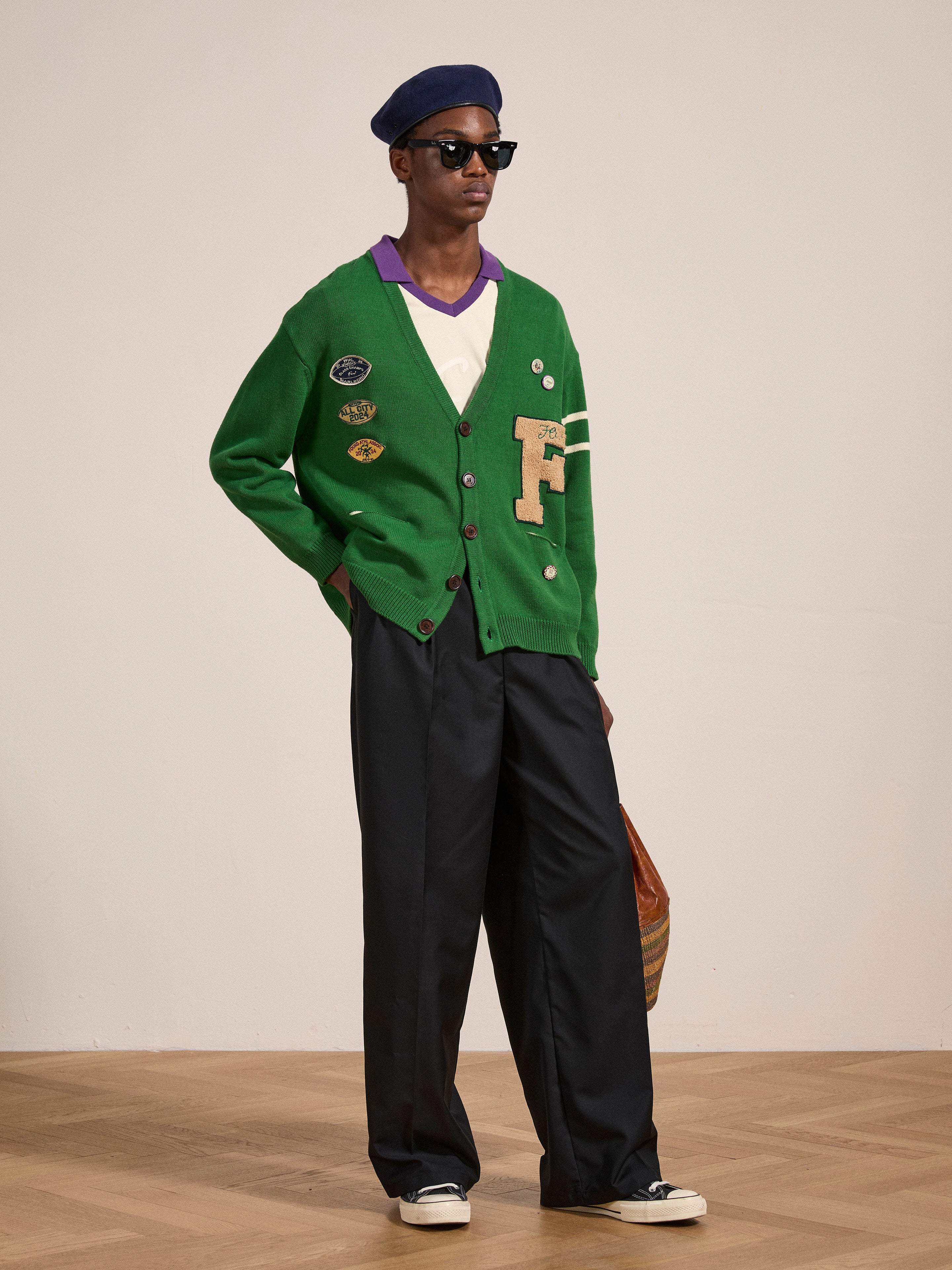 A person wearing a FOUND Pineneedle Varsity Knit Cardigan adorned with a large letter "F," paired with dark wide-leg pants, a black beret, sunglasses, and white shoes, stands indoors on a wooden floor, effortlessly showcasing their vintage look.