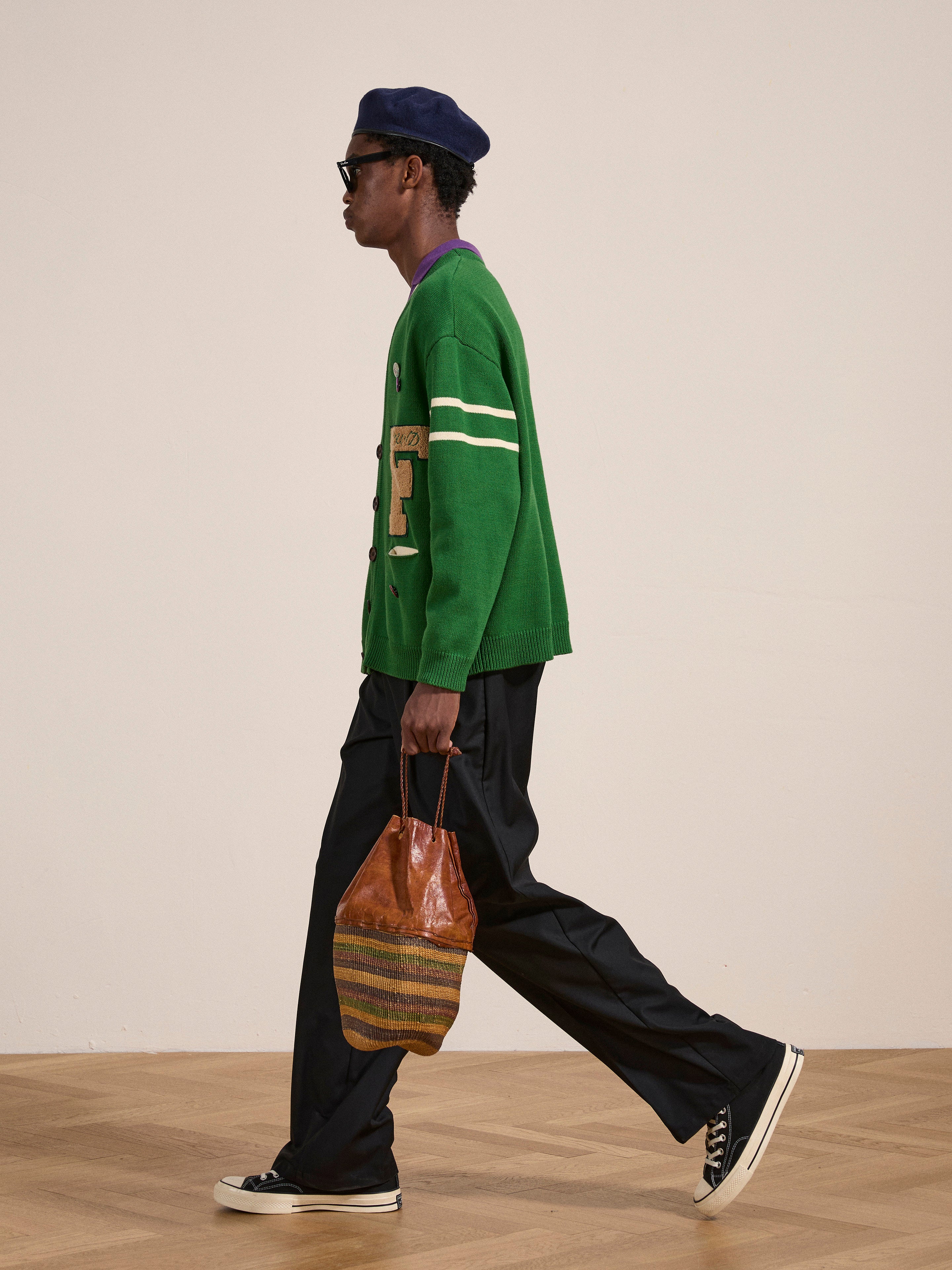 A person wearing a green FOUND Pineneedle Varsity Knit Cardigan, dark pants, and sneakers walks while holding a brown striped bag. They are also wearing a dark beret and sunglasses, giving off a vintage look.