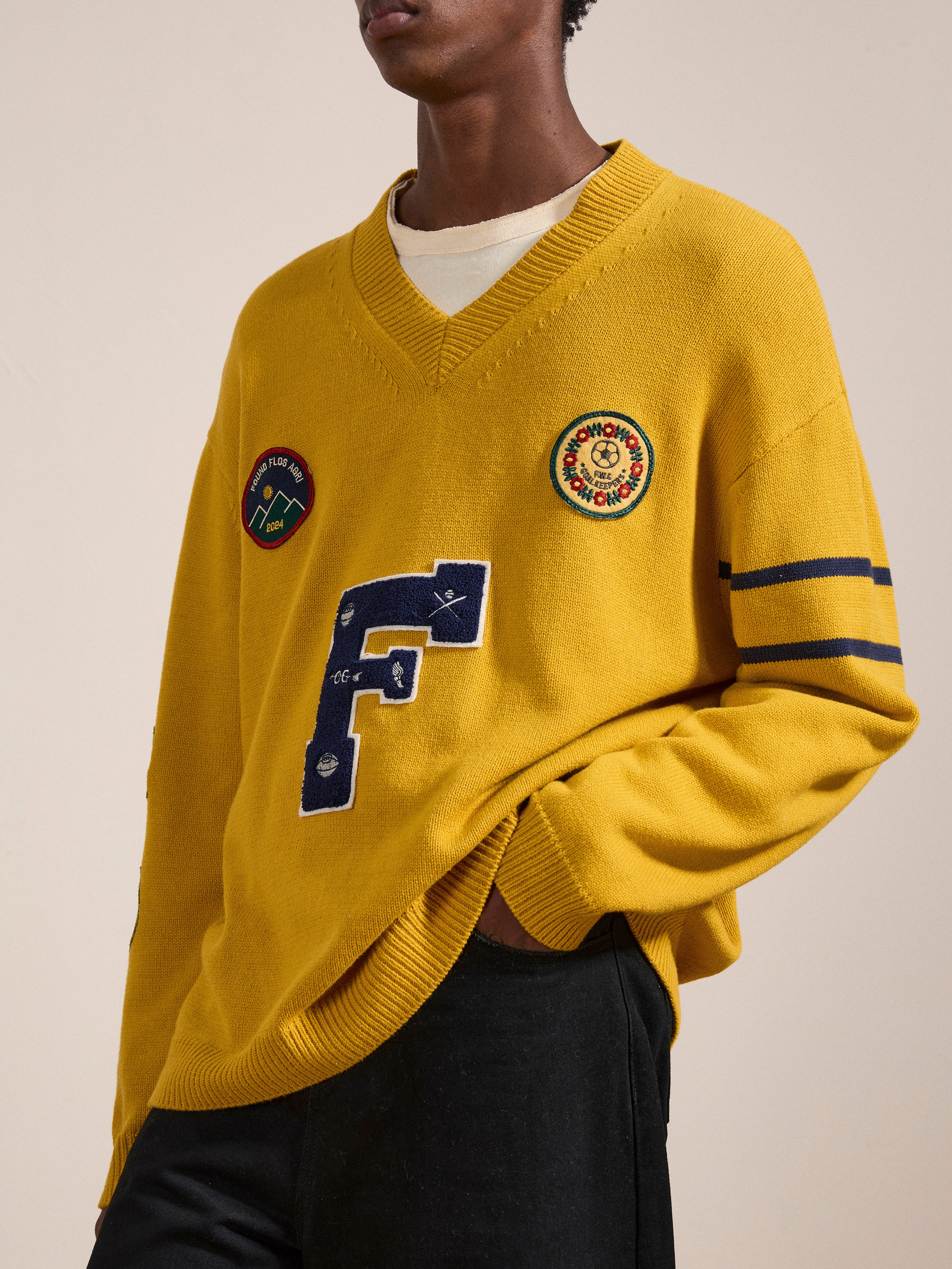 A person wearing a yellow Arrow-Wood Varsity Knit Sweater with a large letter "F" and various patches, paired with black pants, stands against a plain background, showcasing an Ivy League wardrobe with a vintage look from the brand FOUND.