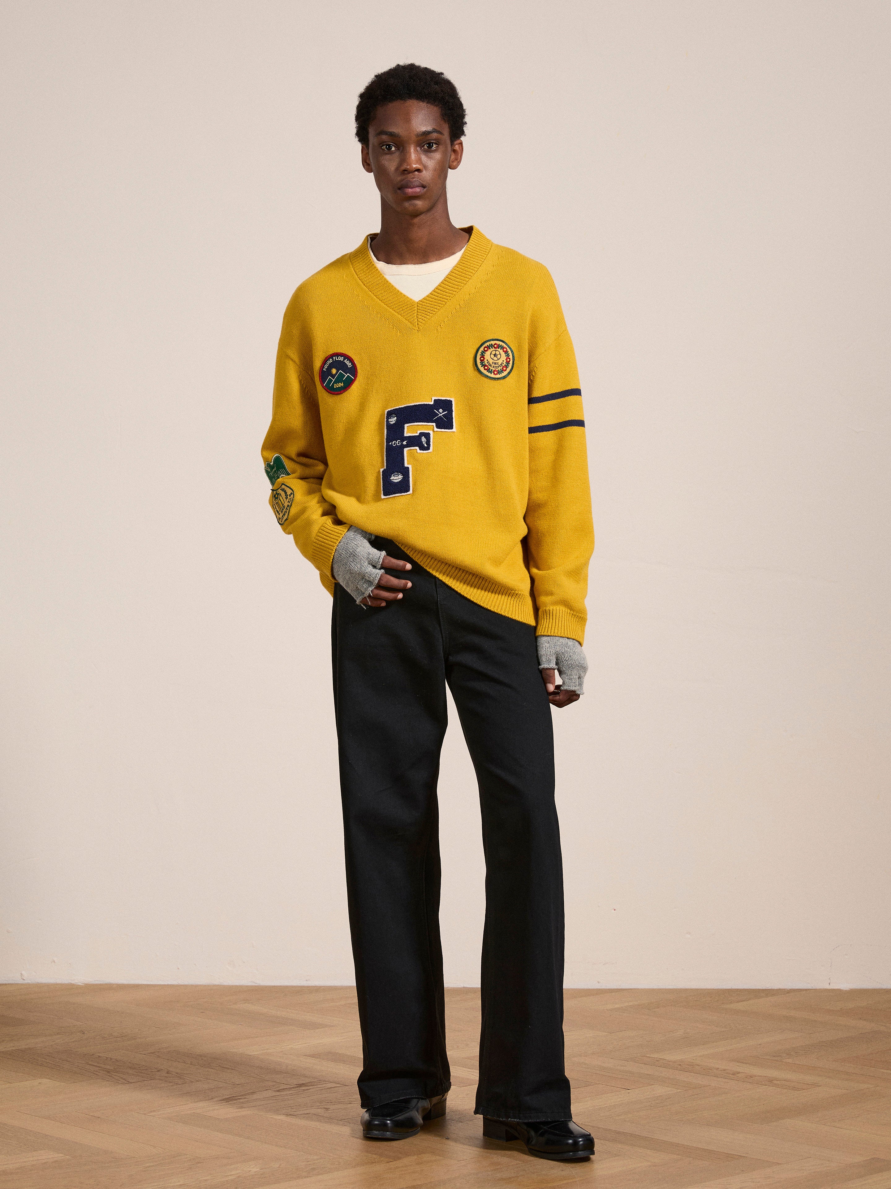 A person stands indoors on a wooden floor, sporting the yellow Arrow-Wood Varsity Knit Sweater by FOUND with patches and black pants. They are also wearing gray fingerless gloves, creating a vintage look.