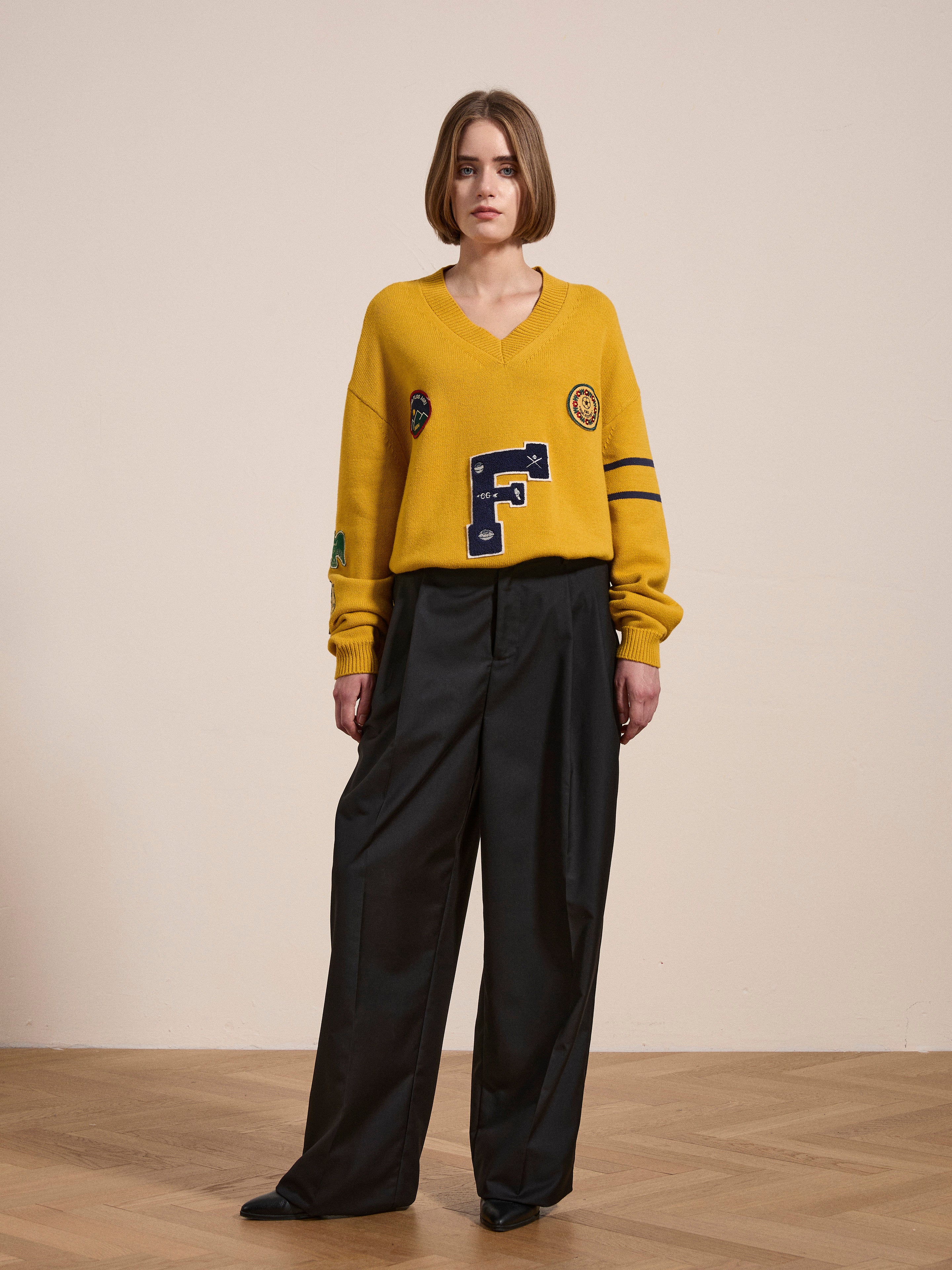 A person stands against a plain backdrop wearing an Arrow-Wood Varsity Knit Sweater by FOUND, featuring letter and patch designs. They pair it with black wide-leg trousers, channeling a vintage look reminiscent of an Ivy League wardrobe.