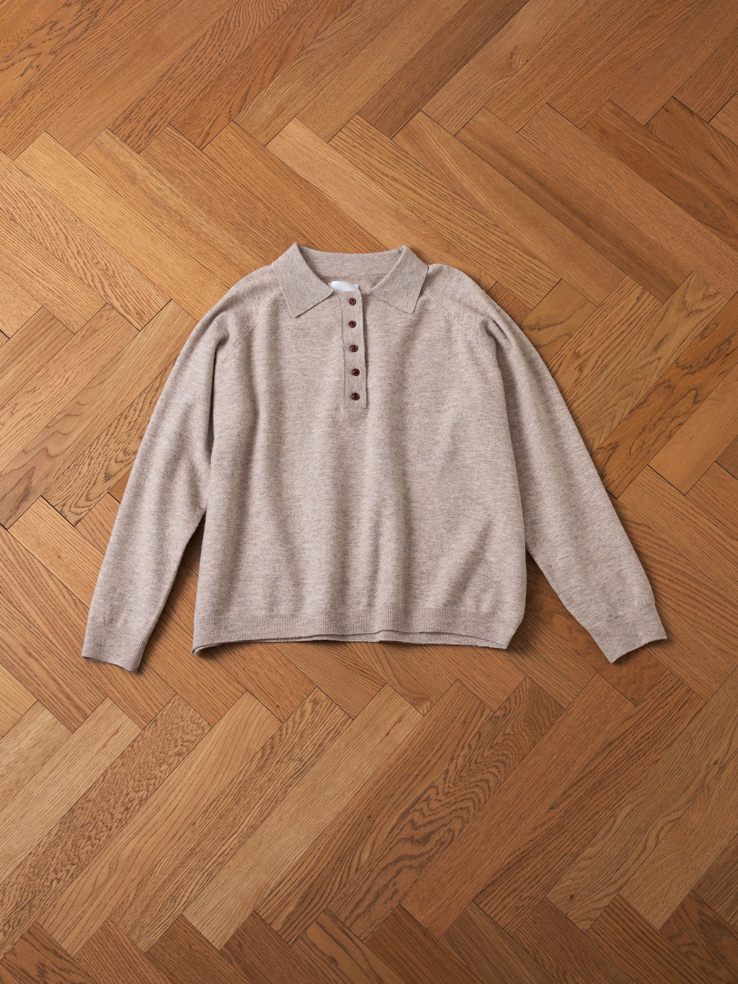 The Hay Collared Knit Henley by FOUND, a beige long-sleeve polo shirt with wooden buttons, lies flat on a wooden herringbone-pattern floor.