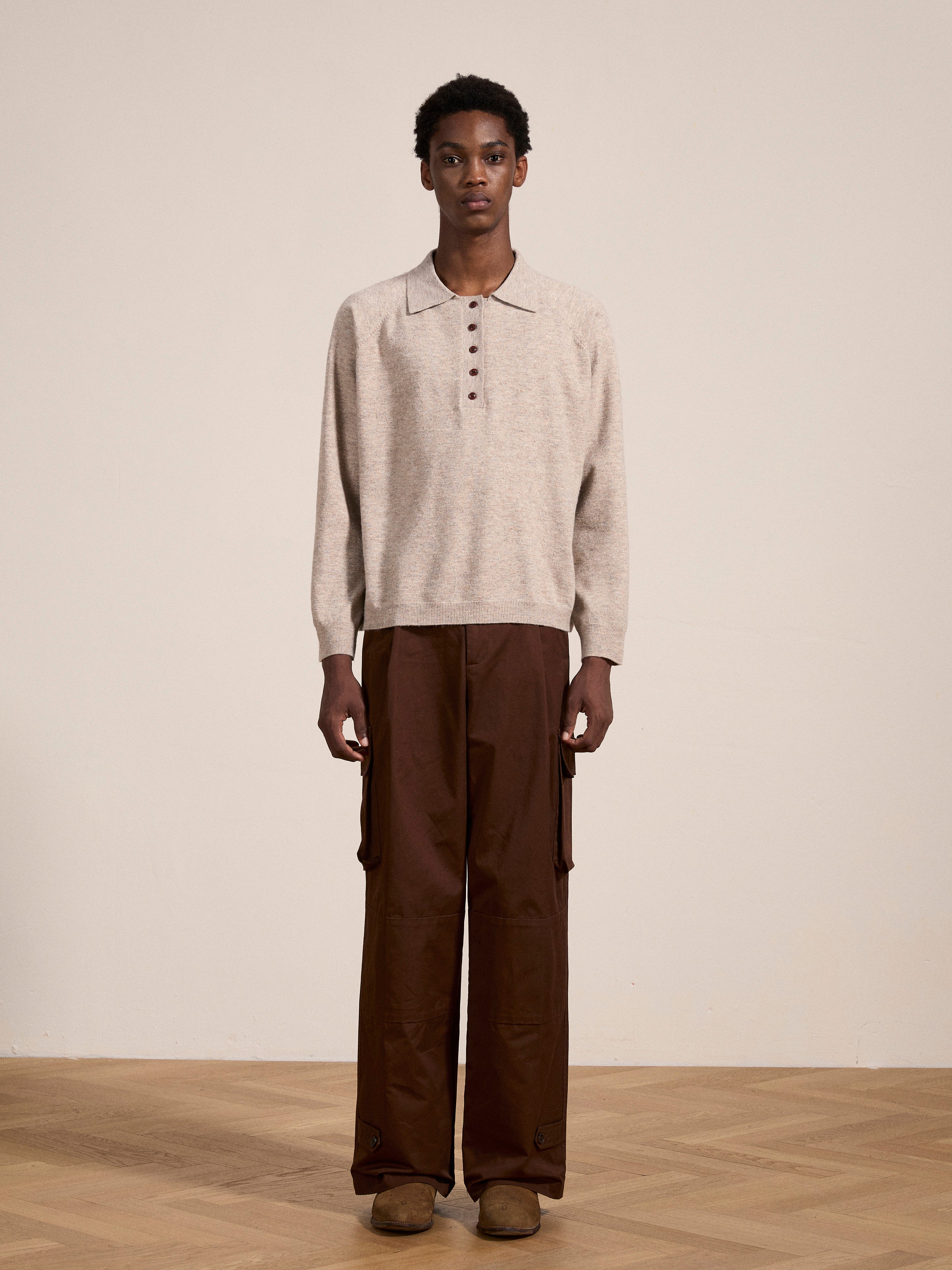 A person stands indoors on a wooden floor, wearing a beige long-sleeve Hay Collared Knit Henley with wooden buttons by FOUND, brown wide-leg cargo pants, and brown shoes. The wall behind the person is plain and off-white.