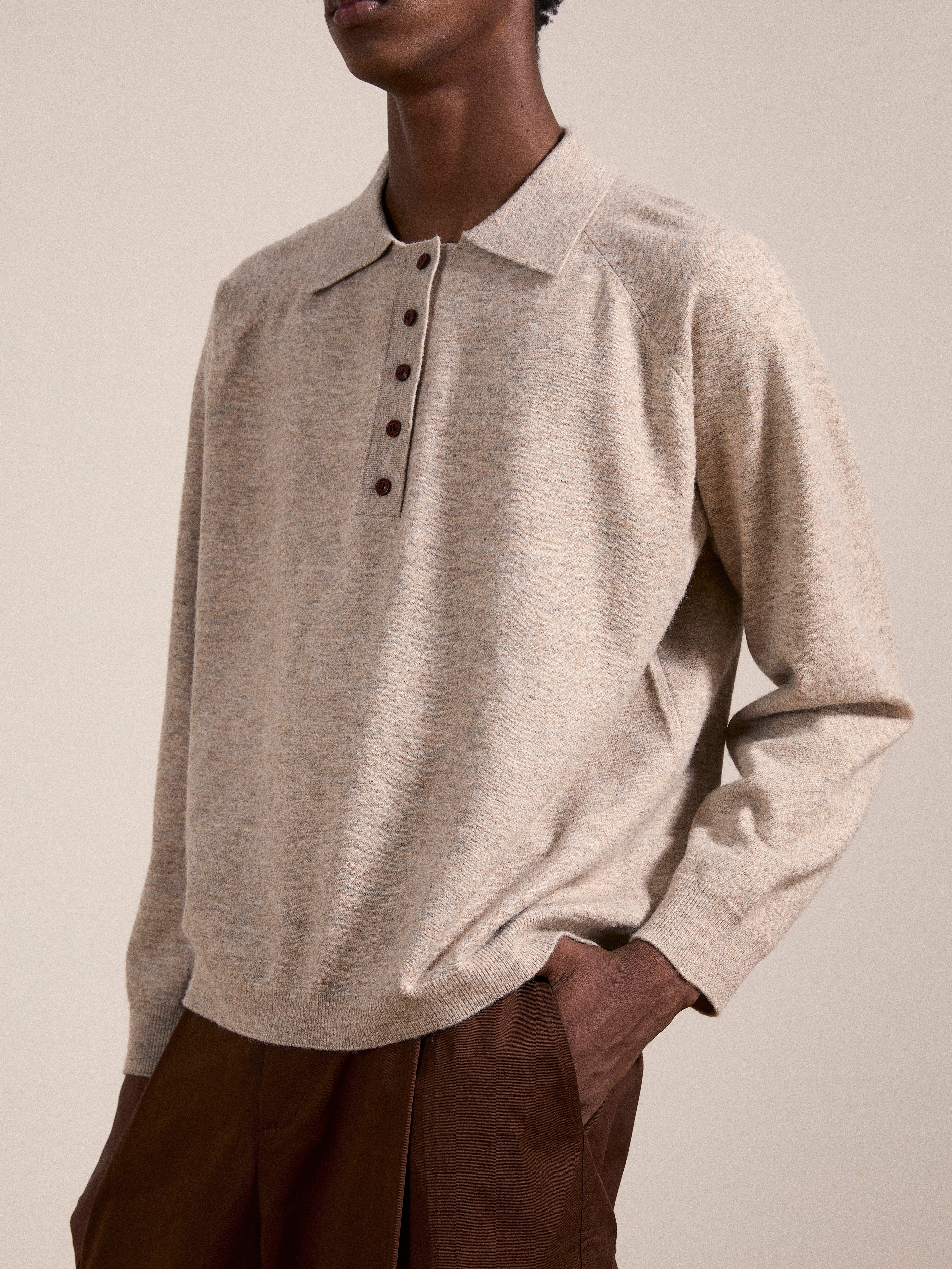 A person wearing a beige, long-sleeved FOUND Hay Collared Knit Henley sweater with wooden buttons and brown trousers, standing with their hands in their pockets. The background is plain and neutral.