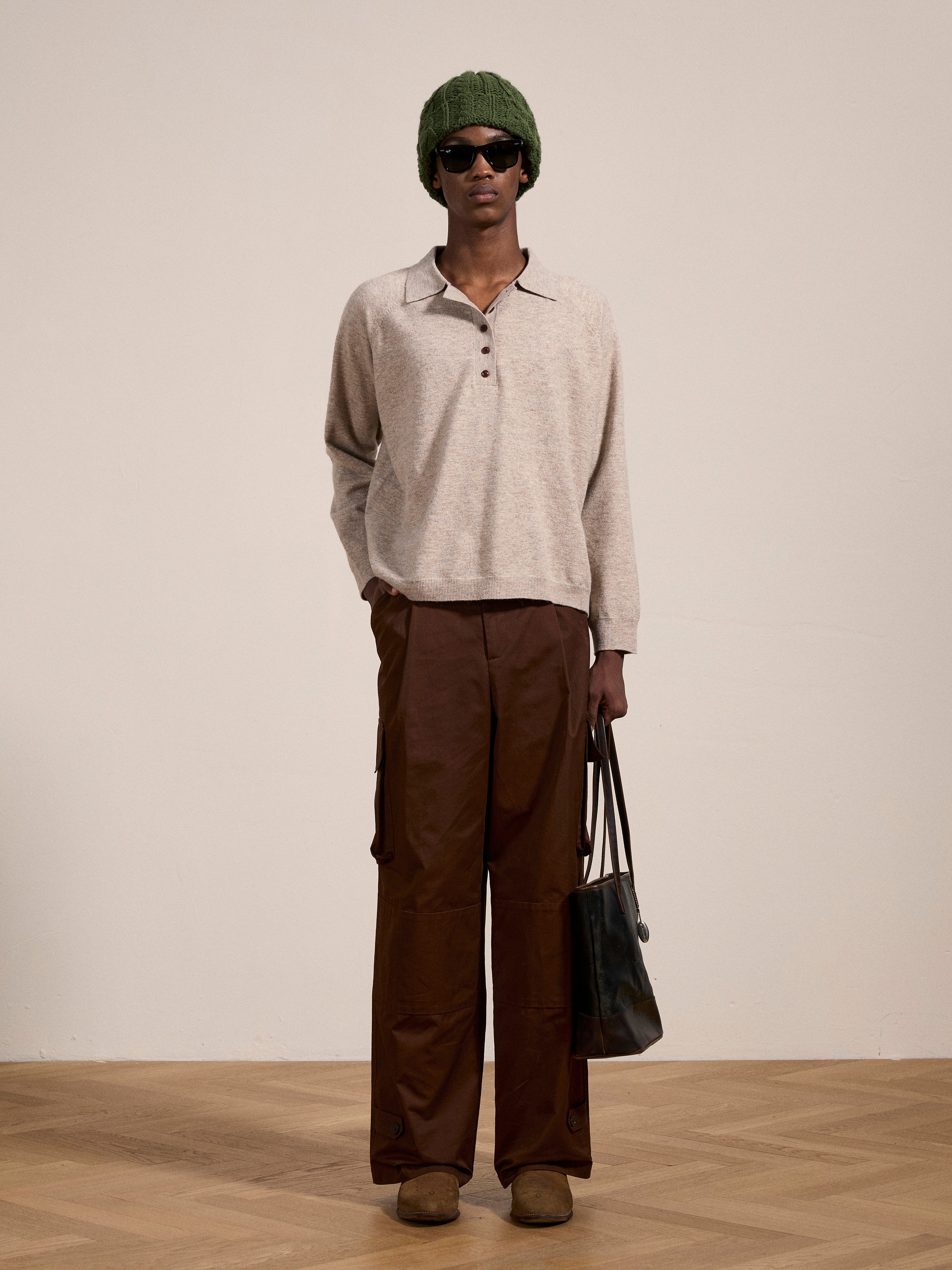 Person stands against a plain wall wearing a green beanie, dark sunglasses, a beige Hay Collared Knit Henley with wooden buttons from FOUND, brown pants, and brown shoes, holding a black tote bag.