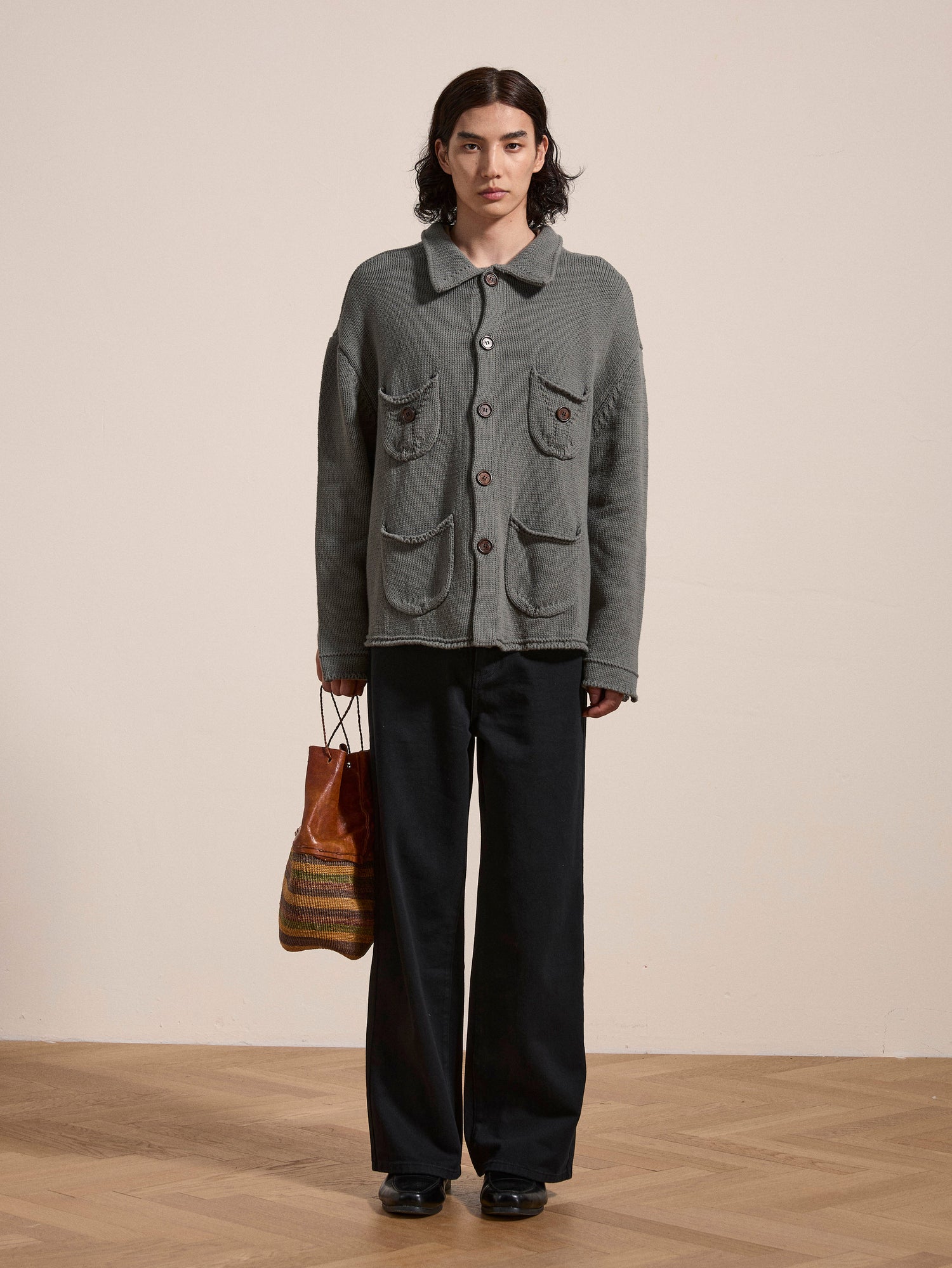 A person wearing the Hunter Knit Collared Knit Cardigan by FOUND, an oversized gray knit cardigan with large wooden buttons and big pockets, along with black pants and black shoes, standing on a wooden floor and holding a striped bag in one hand.