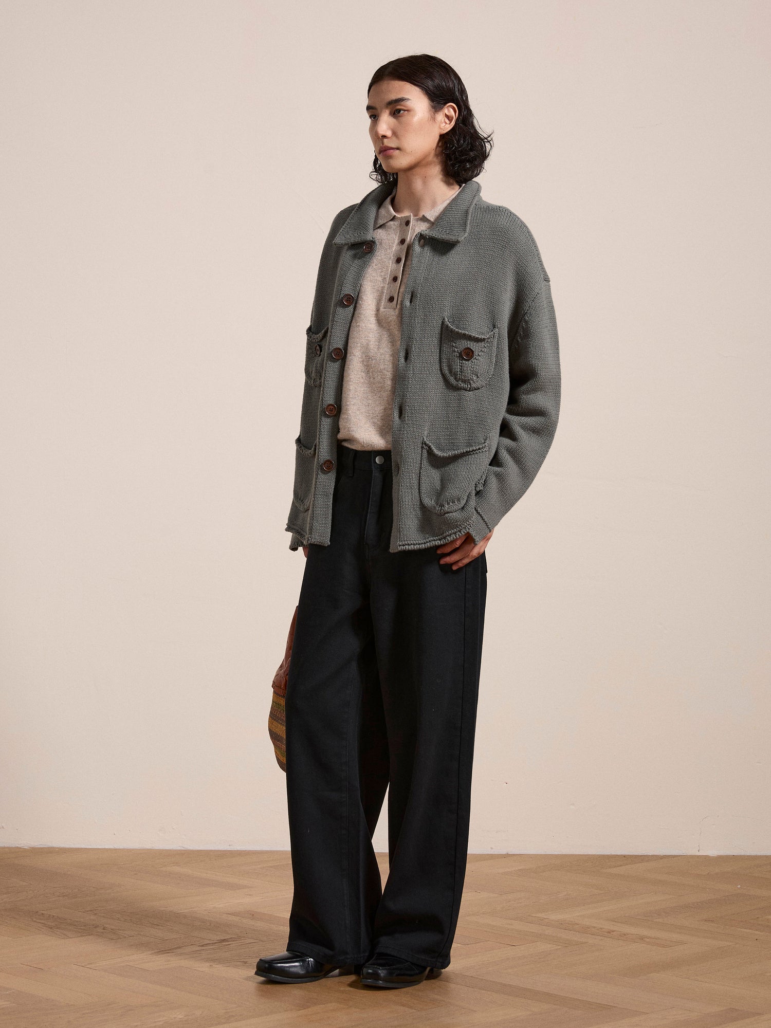 A person stands indoors wearing the FOUND Hunter Knit Collared Knit Cardigan, an oversized grey piece with wooden buttons, paired over a beige shirt and dark wide-legged pants. The floor is wooden, and the background is a plain off-white wall.