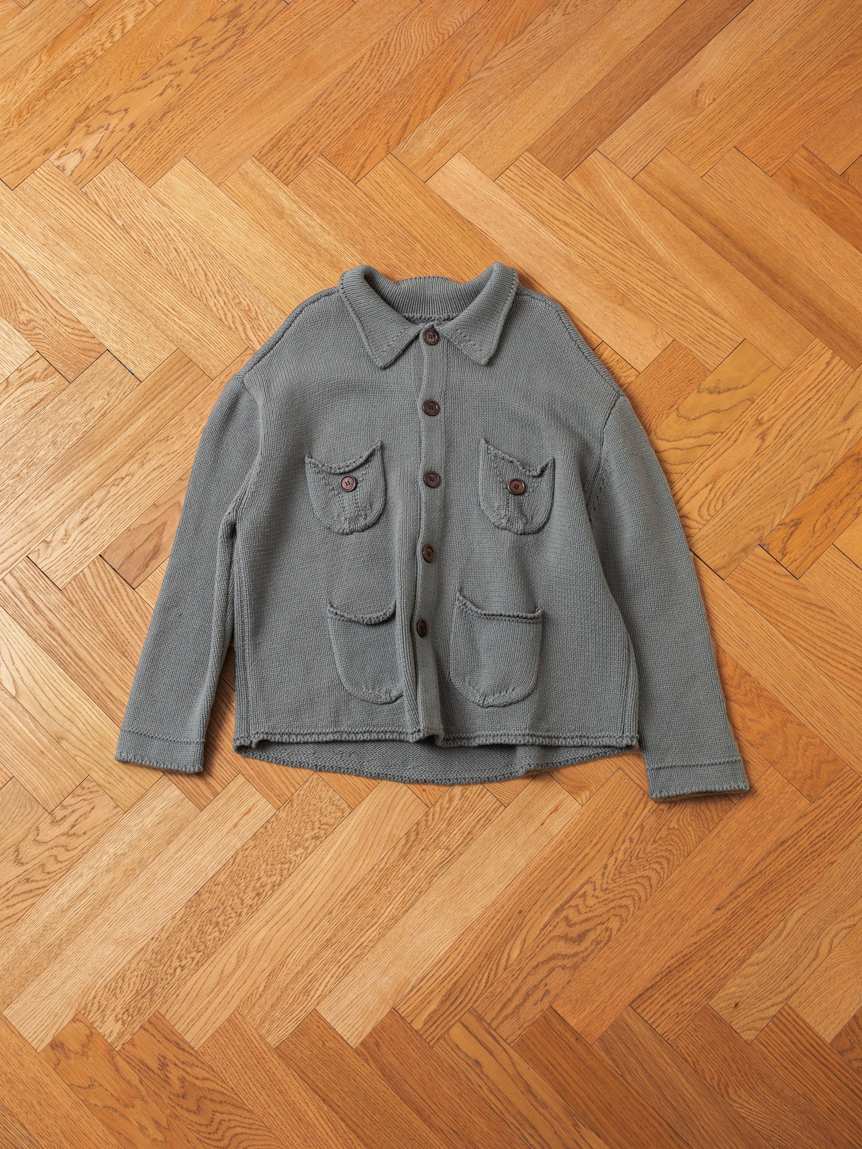 The Hunter Knit Collared Cardigan from FOUND is displayed flat on a wooden herringbone floor, showcasing its gray button-up design with classic wooden buttons, two chest pockets, and two lower pockets.