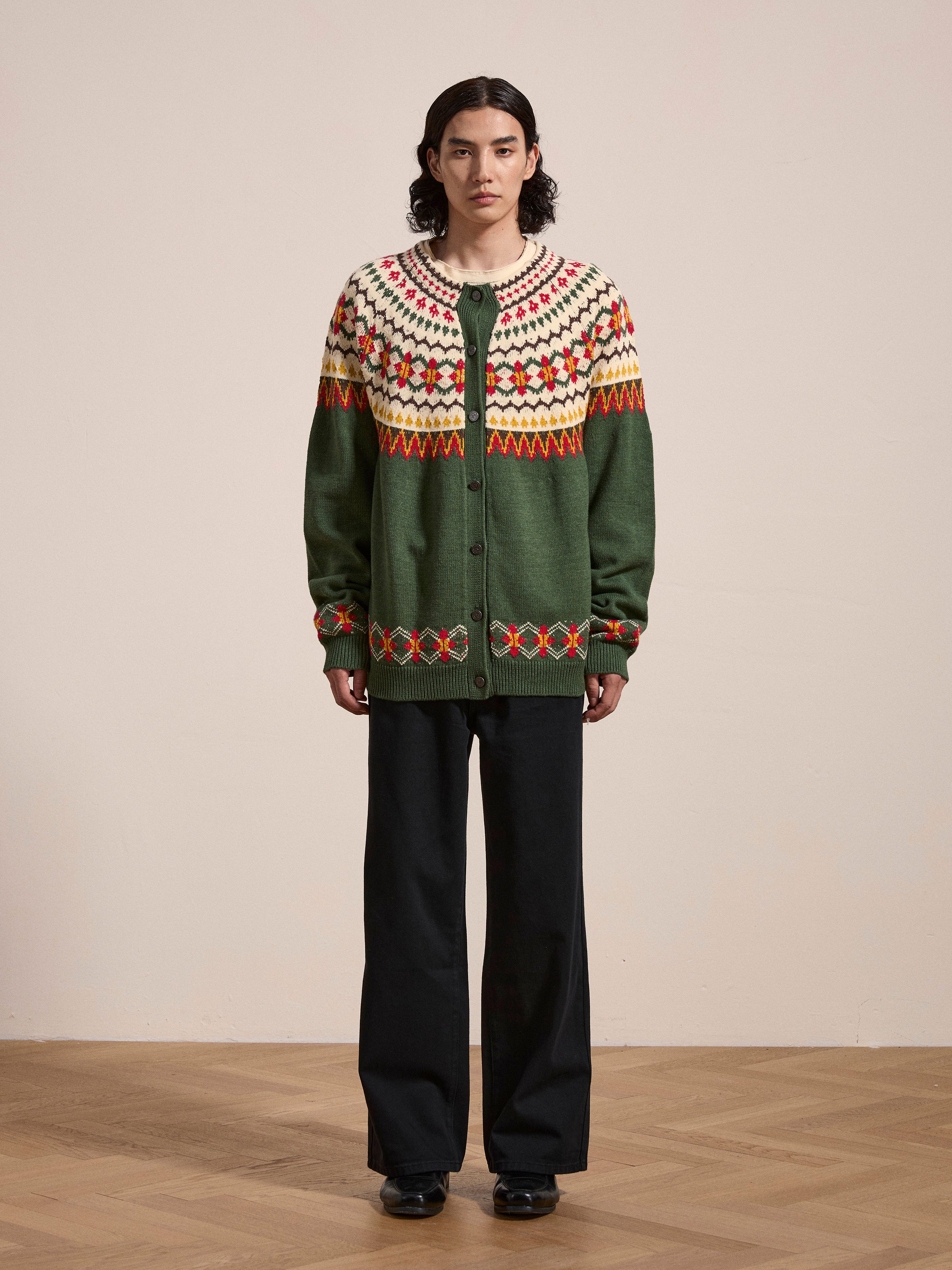 A person standing against a plain background, wearing the FOUND Jahan Isles Knit Cardigan in green, which boasts intricate and colorful Scottish Fair Isle patterns and black pants. The cardigan offers an oversized fit, complemented by antique silver buttons that add a touch of vintage charm to the ensemble.