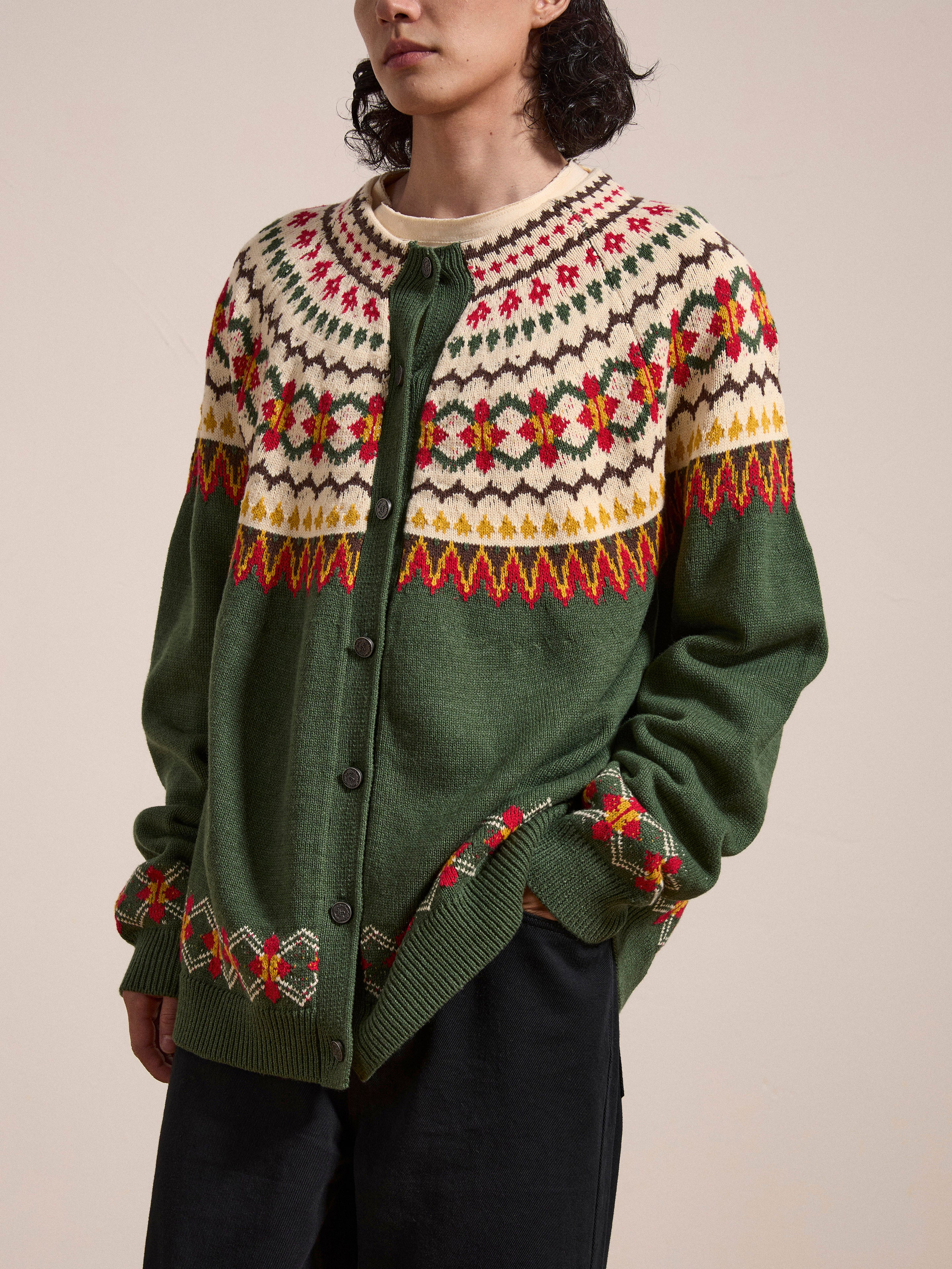 A person wearing the Jahan Isles Knit Cardigan by FOUND, a green, patterned Scottish Fair Isle cardigan with red, white, and yellow designs, featuring antique silver buttons and an oversized fit, stands against a plain, light-colored background.