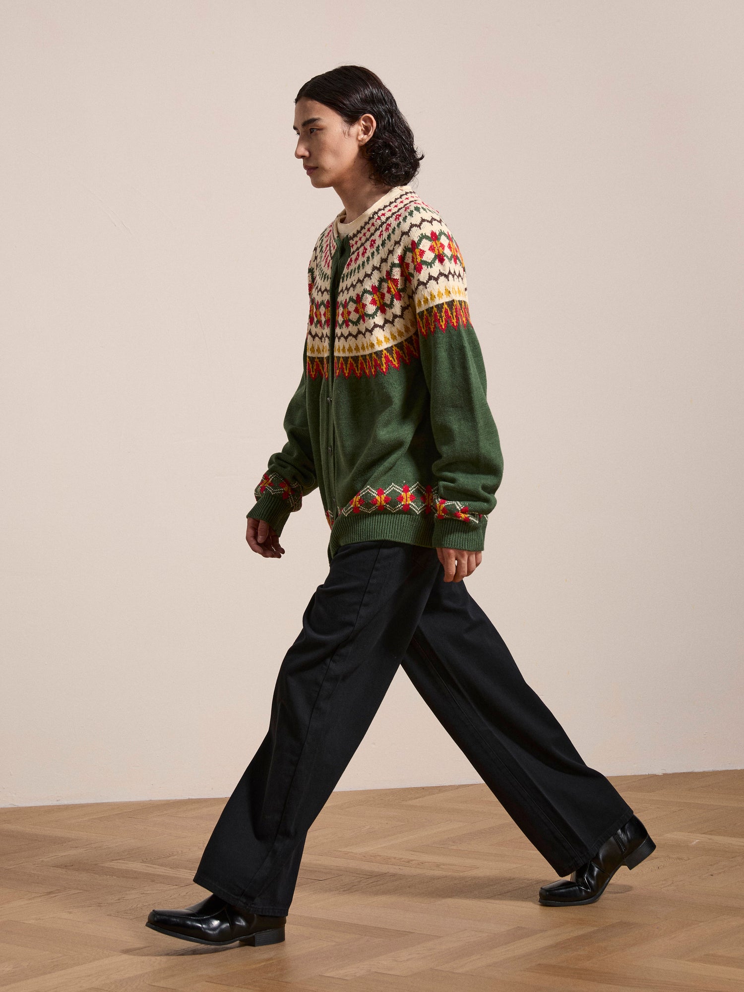 A person with long dark hair wearing the Jahan Isles Knit Cardigan by FOUND, which features an oversized fit with green and white patterns and antique silver buttons, along with black pants and black shoes, walks across a wooden floor against a plain background.