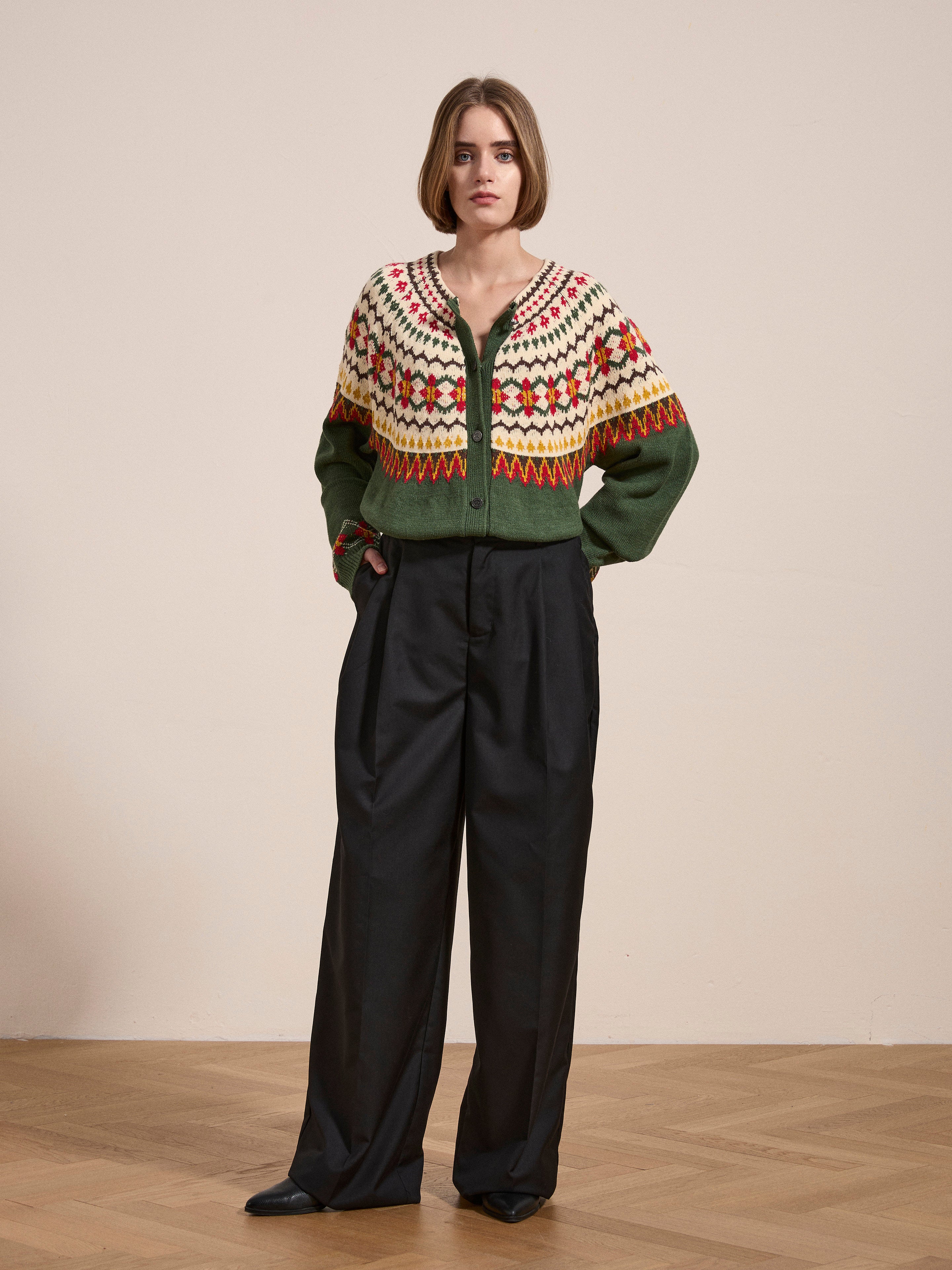 A person stands indoors on a wooden floor, wearing the Jahan Isles Knit Cardigan by FOUND, an oversized green sweater with multicolored patterns, paired with dark wide-leg trousers.