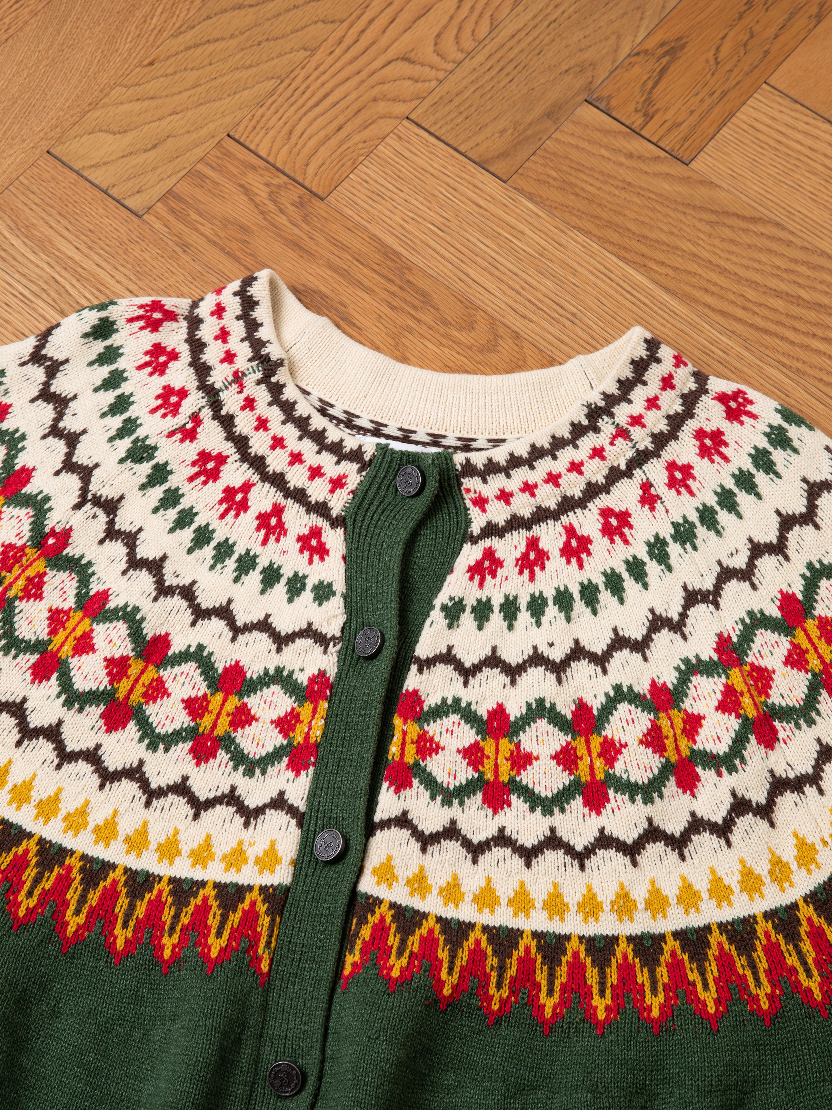 The Jahan Isles Knit Cardigan by FOUND, featuring an oversized Nordic pattern in red, green, yellow, and white with antique silver buttons down the front, is laid out on a wooden floor.