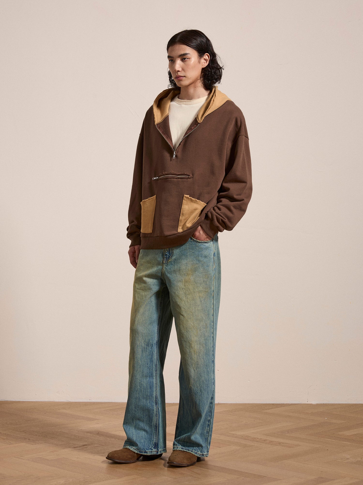 A person in a vintage-feel brown Quarter Zip Two Tone Hoodie by FOUND, paired with light blue wide-leg jeans, stands on a wooden floor against a plain wall.