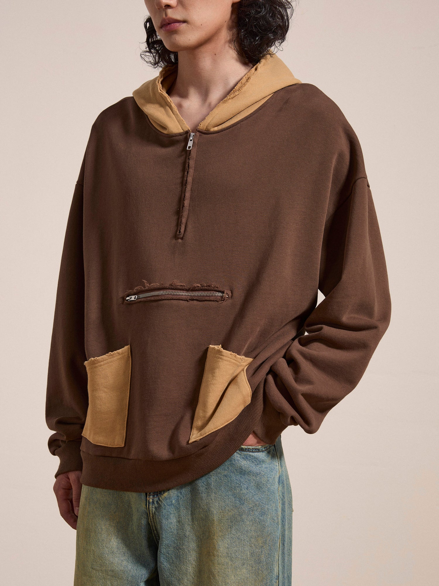 A person wearing the FOUND Quarter Zip Two Tone Hoodie, featuring a brown base with beige accents and a convenient zipper pocket, paired with light blue jeans, exuding a vintage feel.