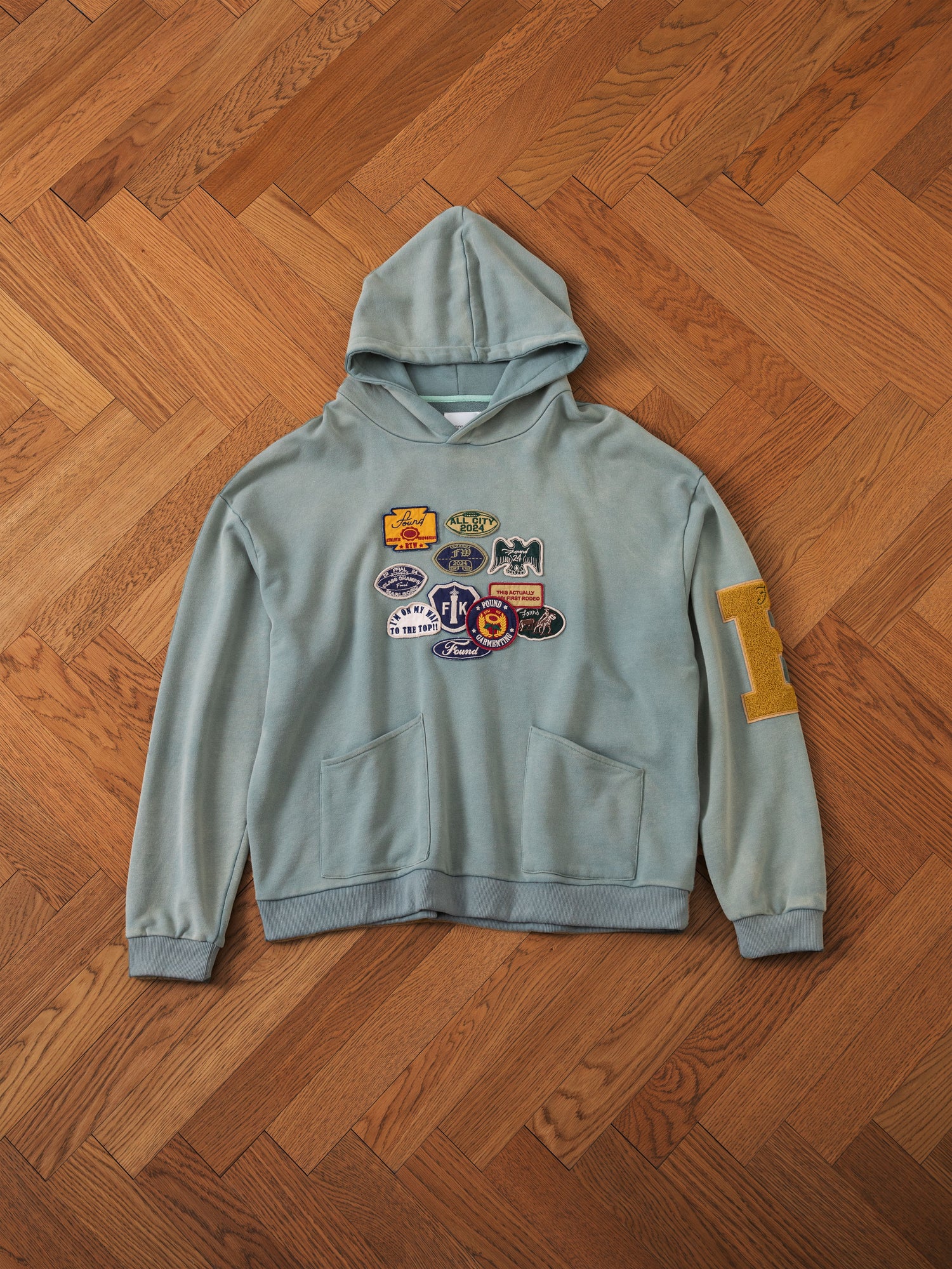 A light green Multi Patch Hoodie from FOUND, crafted from French Terry cotton and adorned with multiple colorful patches on the front and a large yellow 