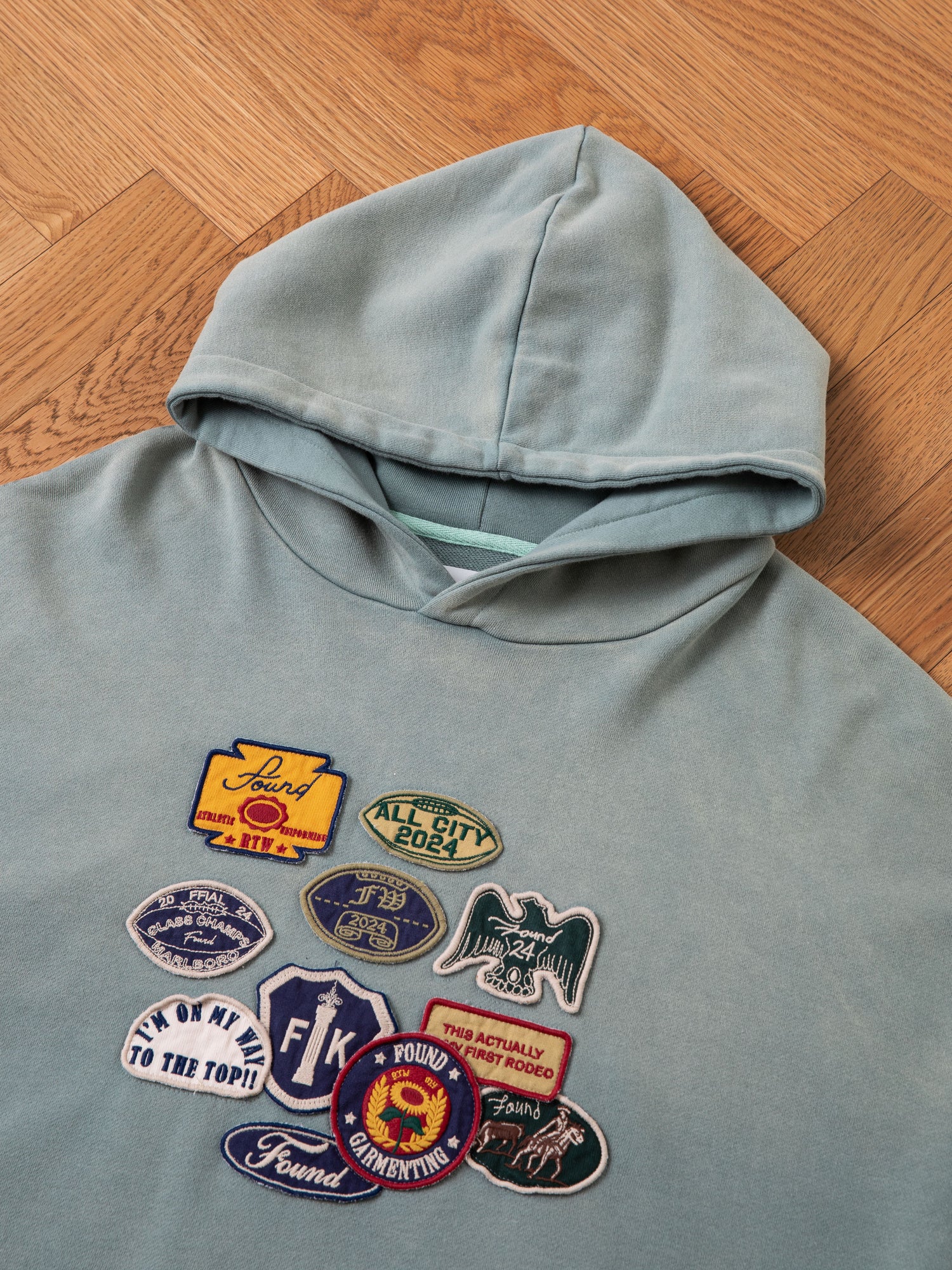 A Multi Patch Hoodie by FOUND, crafted from French Terry cotton, adorned with an array of colorful vintage-inspired embroidered patches on the front, is laid flat on a wooden herringbone parquet floor.