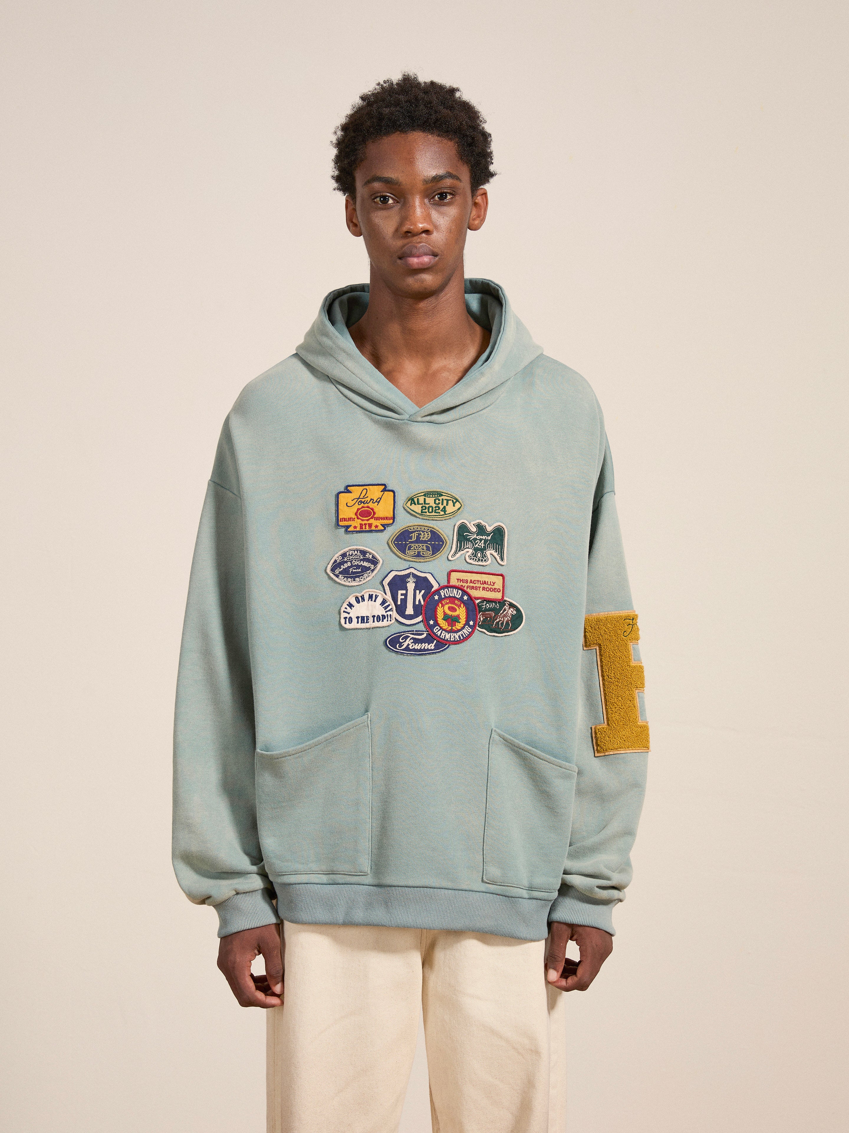 A person stands wearing a Multi Patch Hoodie by FOUND, a light blue, vintage-inspired sweatshirt adorned with multiple colorful patches on the front and featuring a large letter "E" on the left sleeve. Made from French Terry cotton, they have a neutral expression and wear light-colored pants.