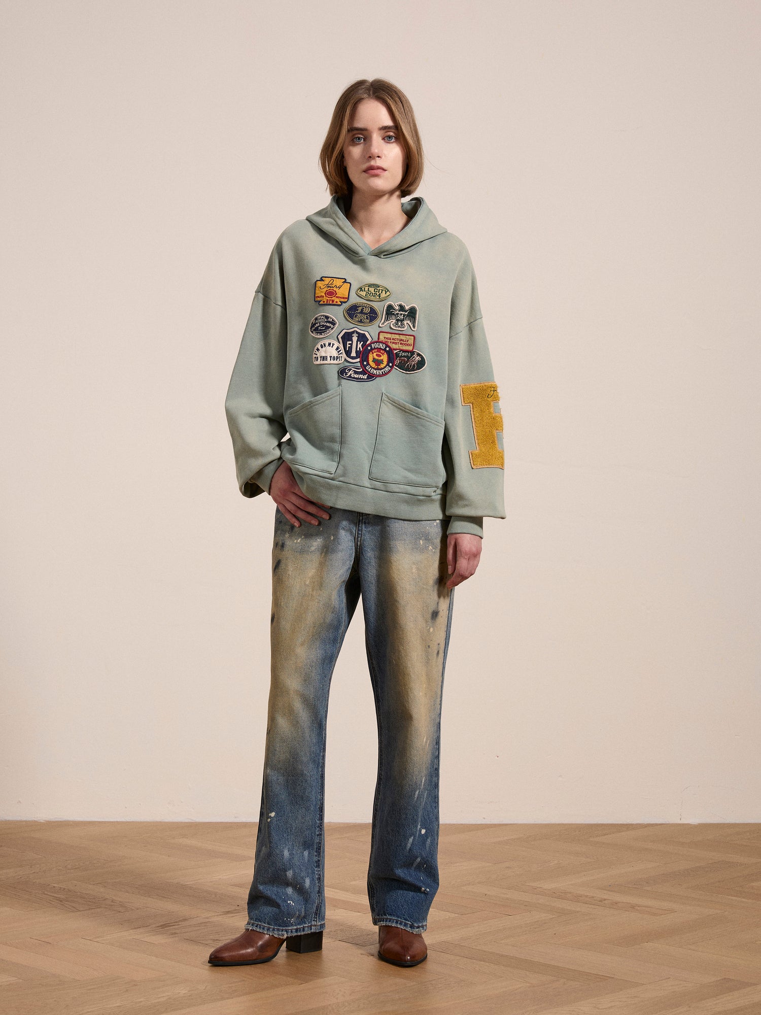 A person is standing indoors wearing a FOUND Multi Patch Hoodie made from French Terry cotton, paired with loose-fitting faded jeans and brown shoes. The background is a plain off-white wall, and the floor features a vintage-inspired parquet pattern.