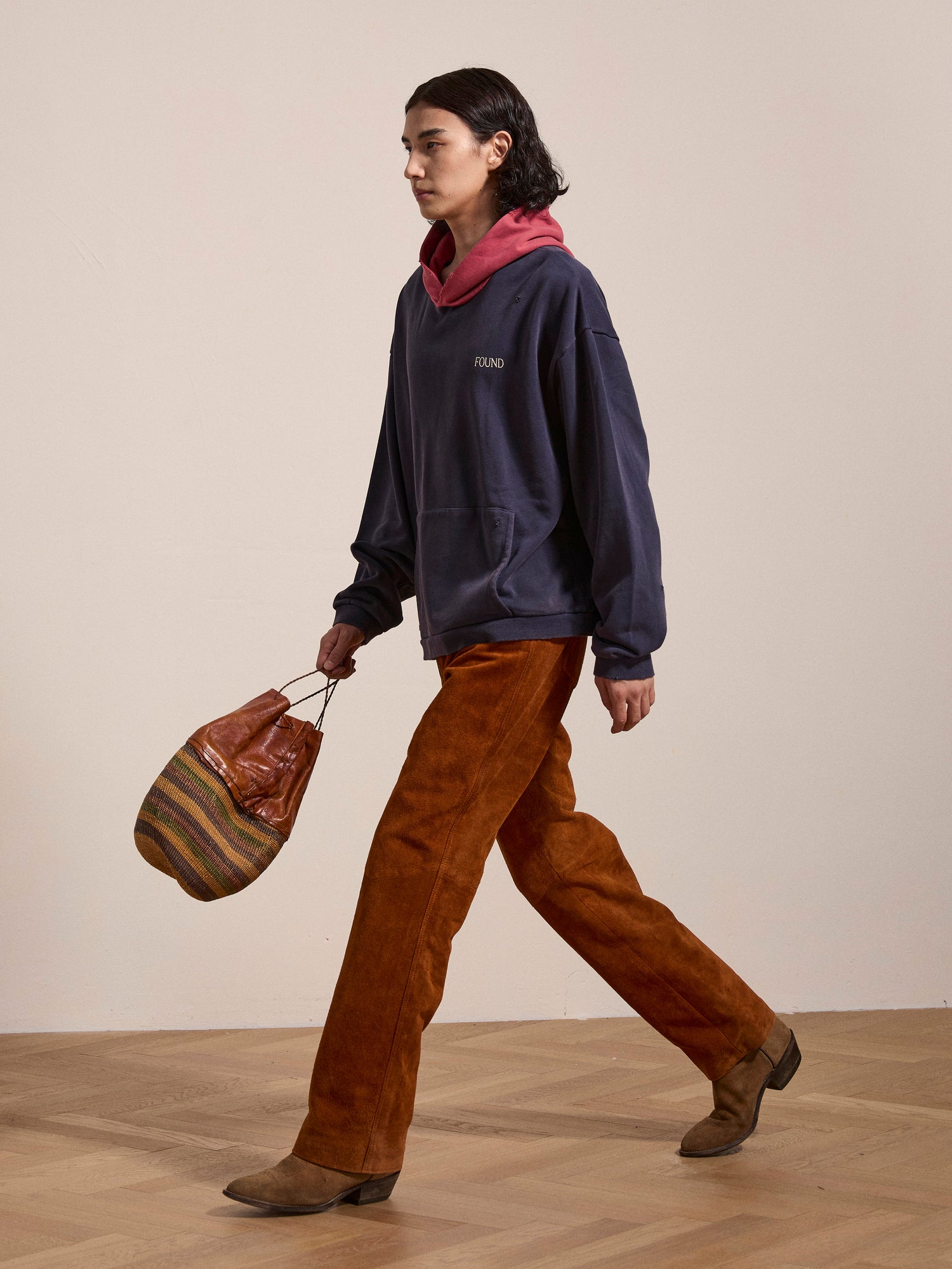 Person walking across a room wearing a dark FOUND Distressed Logo Hoodie, rust-colored pants, and brown shoes, carrying a colorful striped bag.