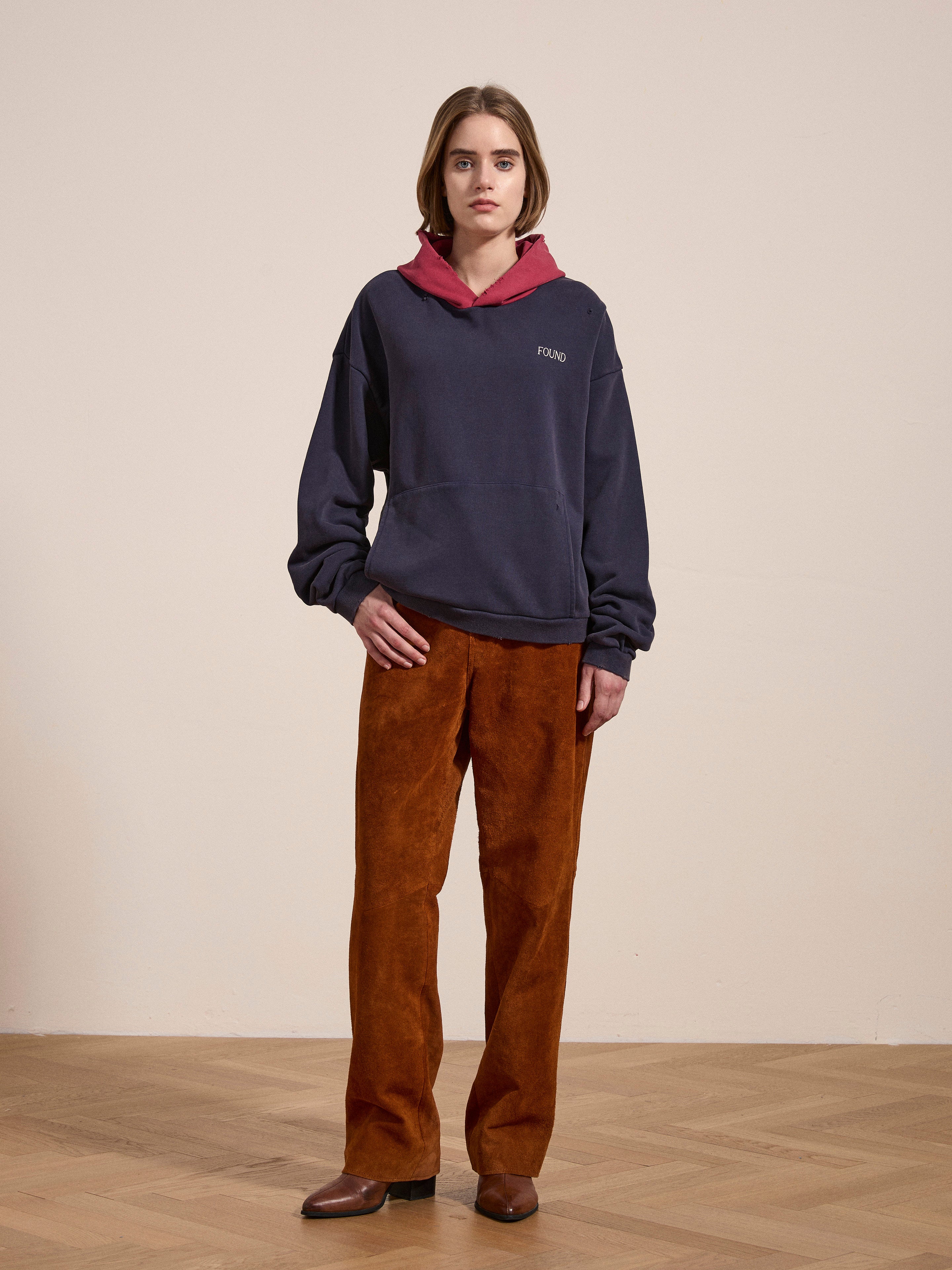 A person stands against a plain background, wearing the FOUND Distressed Logo Hoodie, which is navy with a red hood and drawn strings, showcasing a distressed logo for that vintage feel. They pair it with brown corduroy pants and brown shoes.