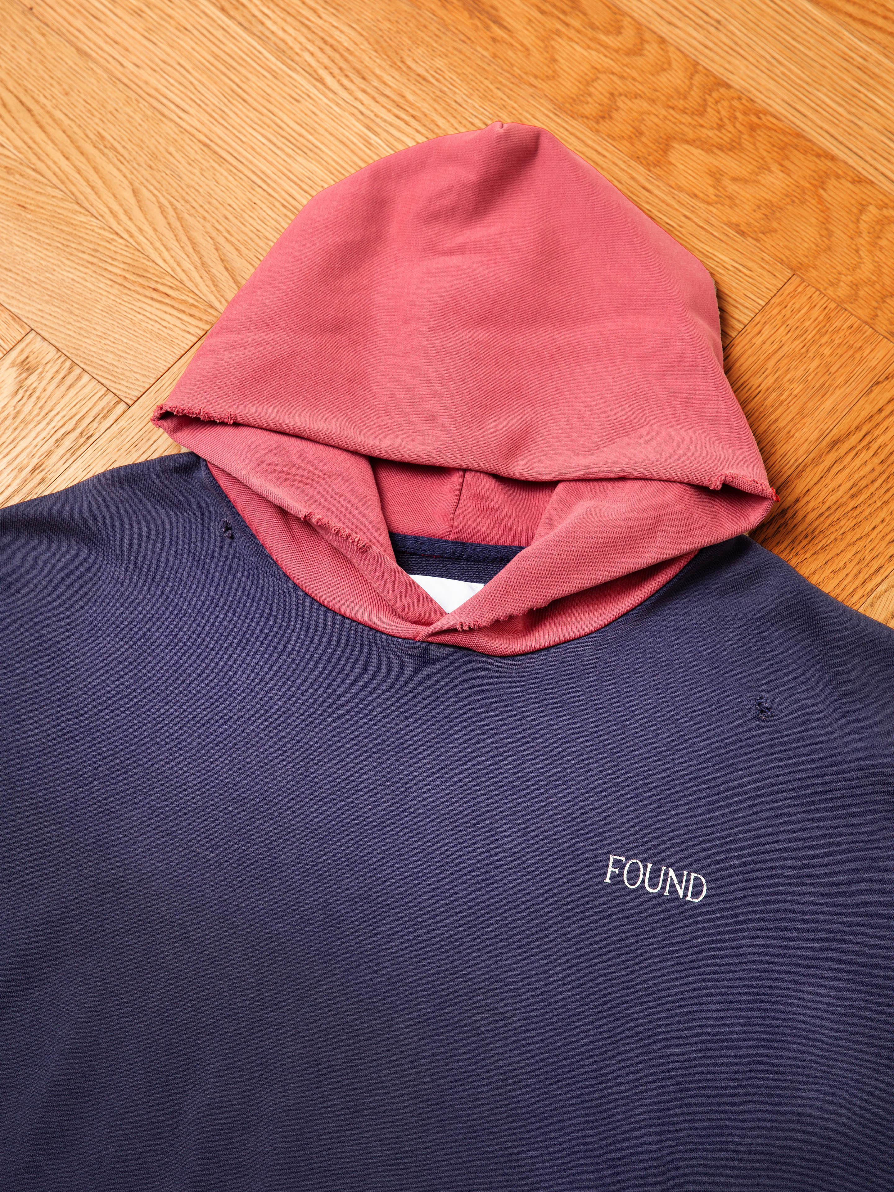 A navy blue Distressed Logo Hoodie from FOUND, featuring a red hood, is laid flat on a wooden floor. Crafted from French terry cotton, this hoodie boasts the word "FOUND" elegantly embroidered in white across the chest for a vintage feel.