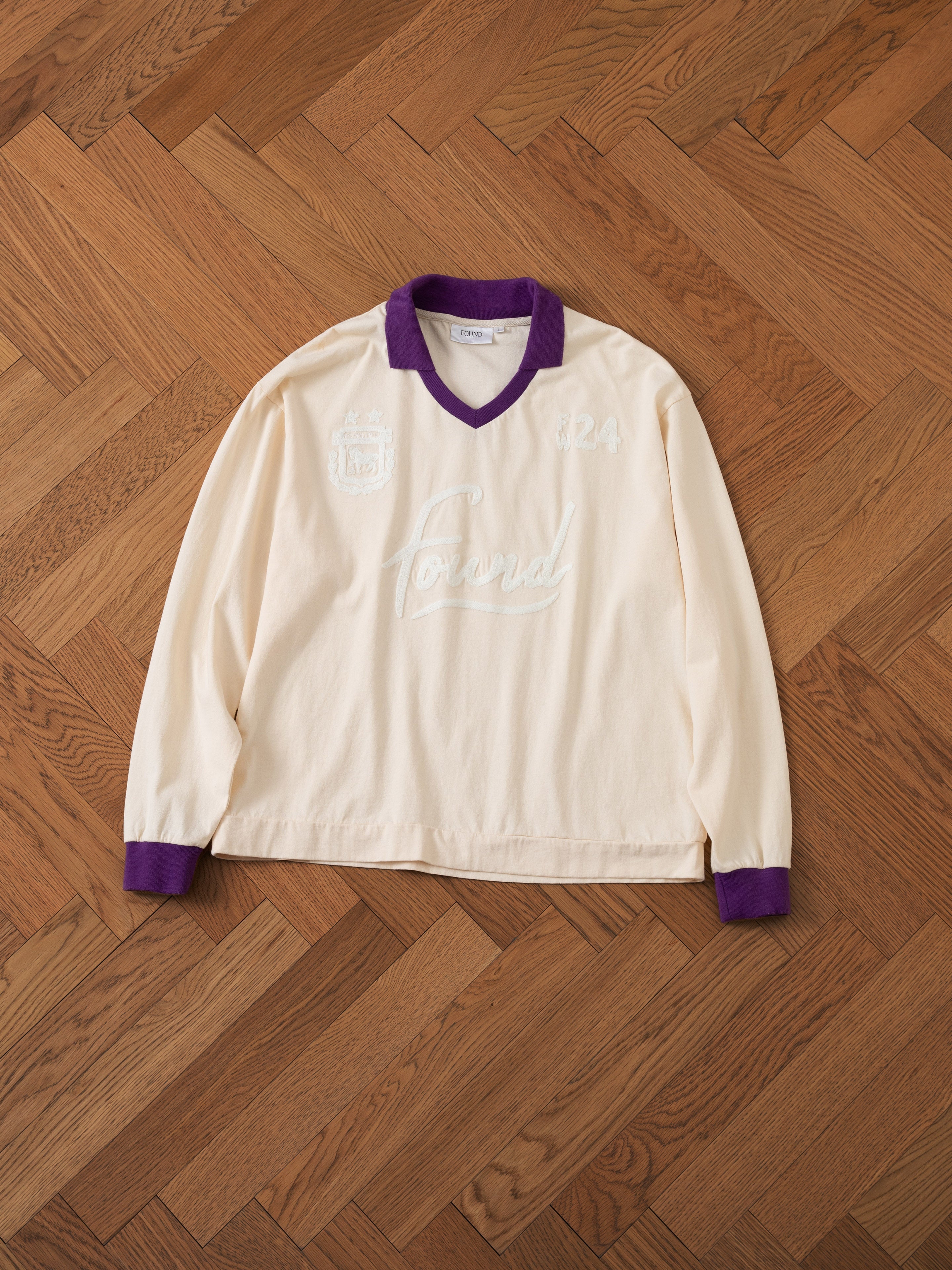 The Sahara Sun Jersey by FOUND, a long-sleeve off-white shirt with purple collar and cuffs reminiscent of vintage athletic uniforms, laid flat on a wooden floor. The shirt features "Forward" and "1924" text along with a crest on the front.