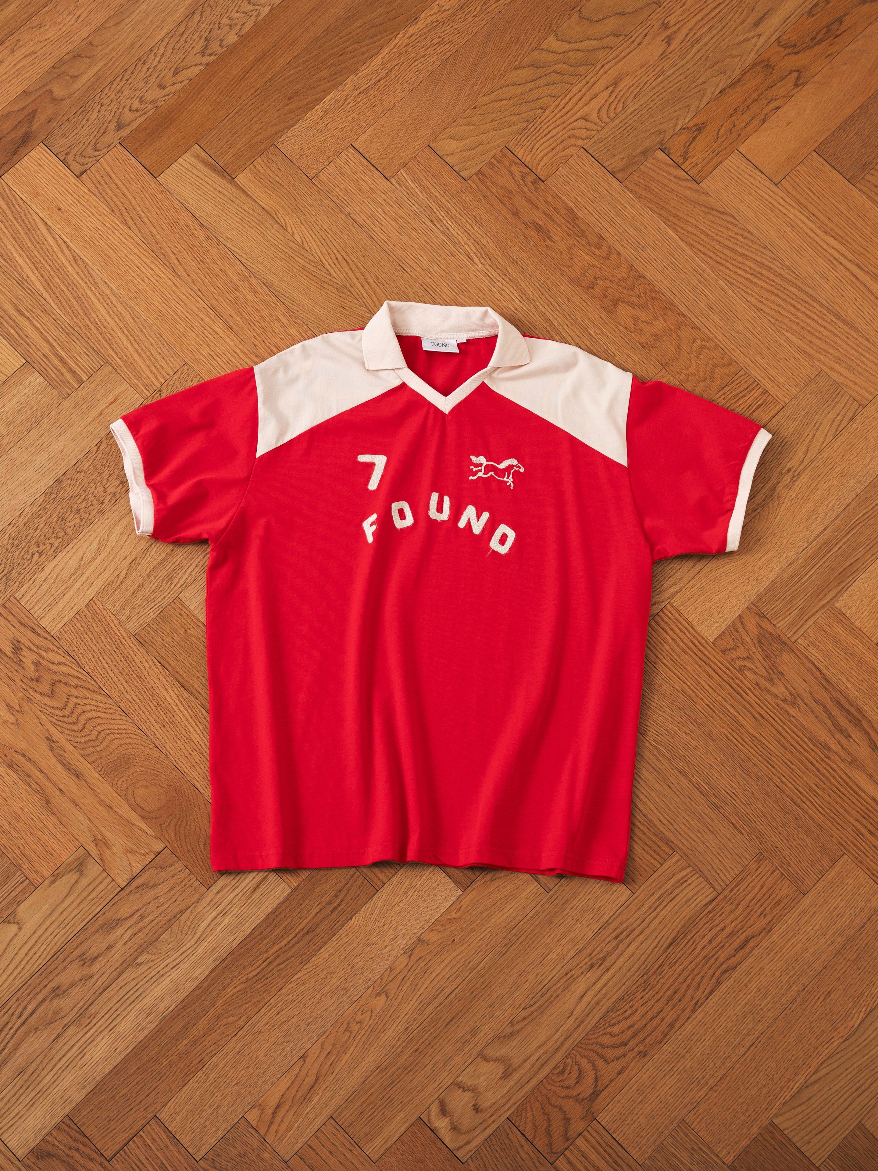 A Goji Red Jersey by FOUND, featuring the number "7" and the word "FOUND" emblazoned on the front along with a contrast color yoke and collar, beautifully displayed on a wooden herringbone floor.