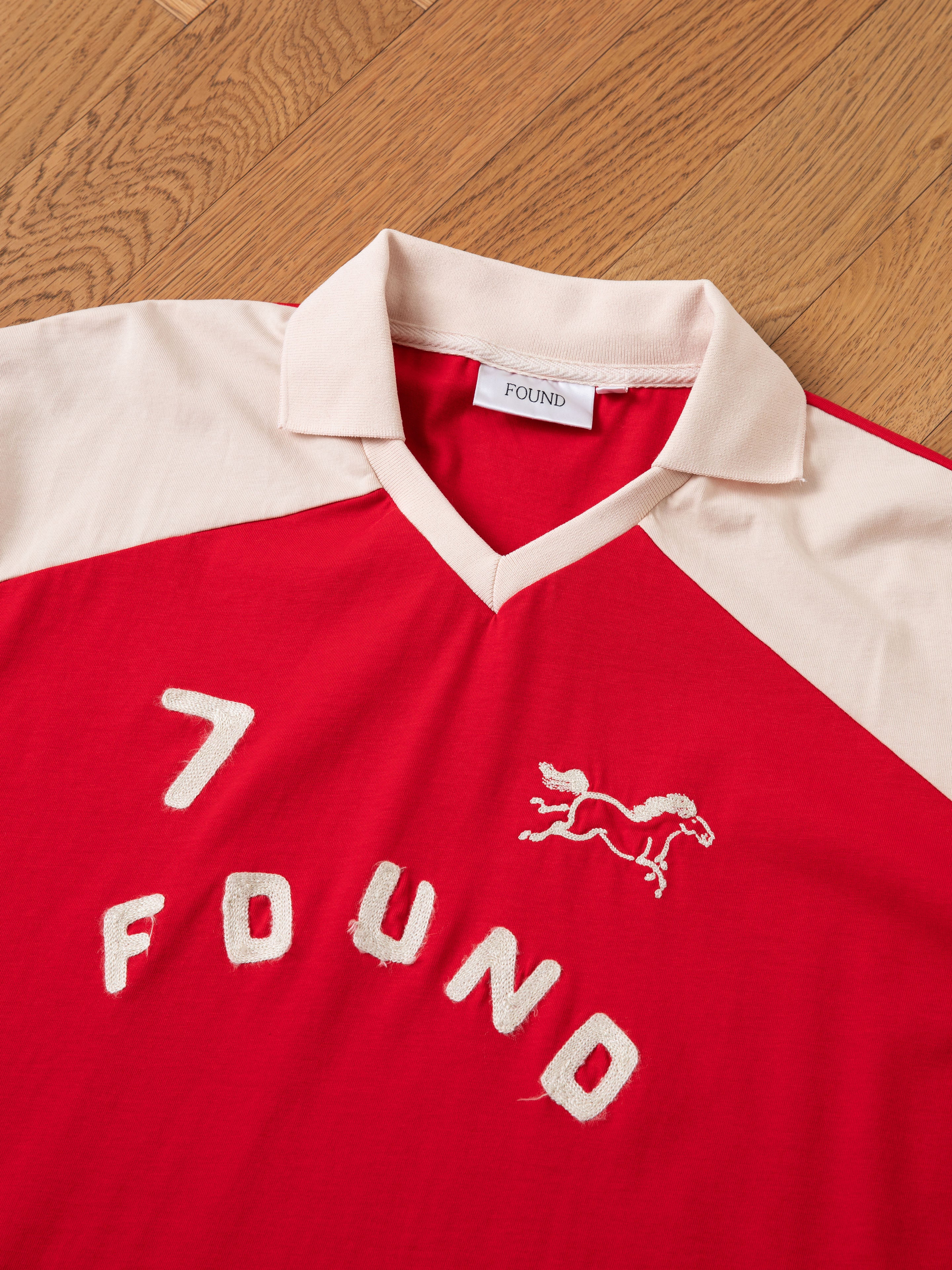 Introducing the Goji Red Jersey by FOUND, a tribute to classic athletic uniforms. This red shirt features white accents and a beige contrast color yoke and collar, displaying the number 7, the brand name 'FOUND,' and an embroidered outline of a horse on the front. The shirt is showcased laid flat on a wood surface.