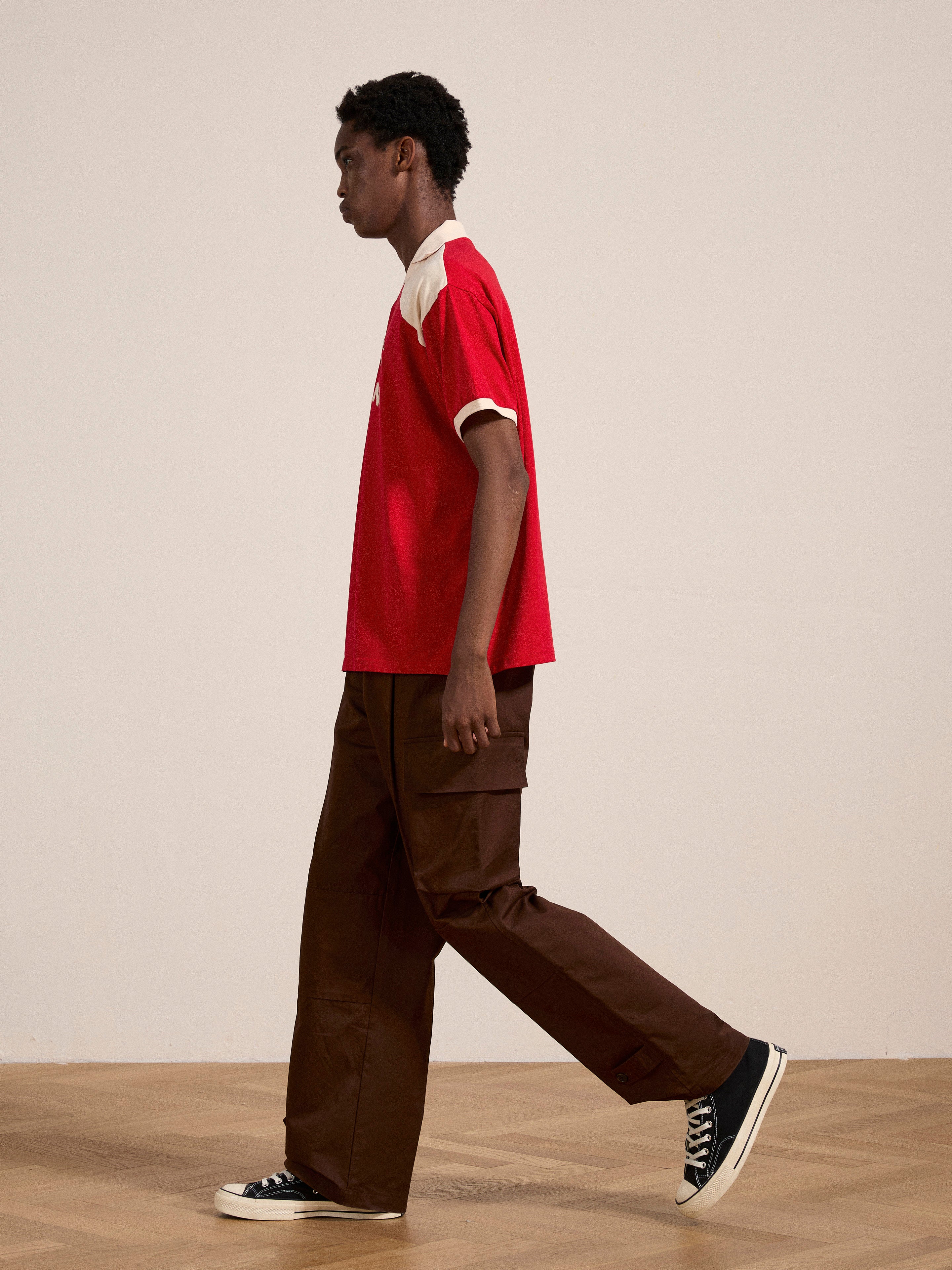 A person in a Goji Red Jersey by FOUND and brown pants walks across a wooden floor against a plain, light-colored background.