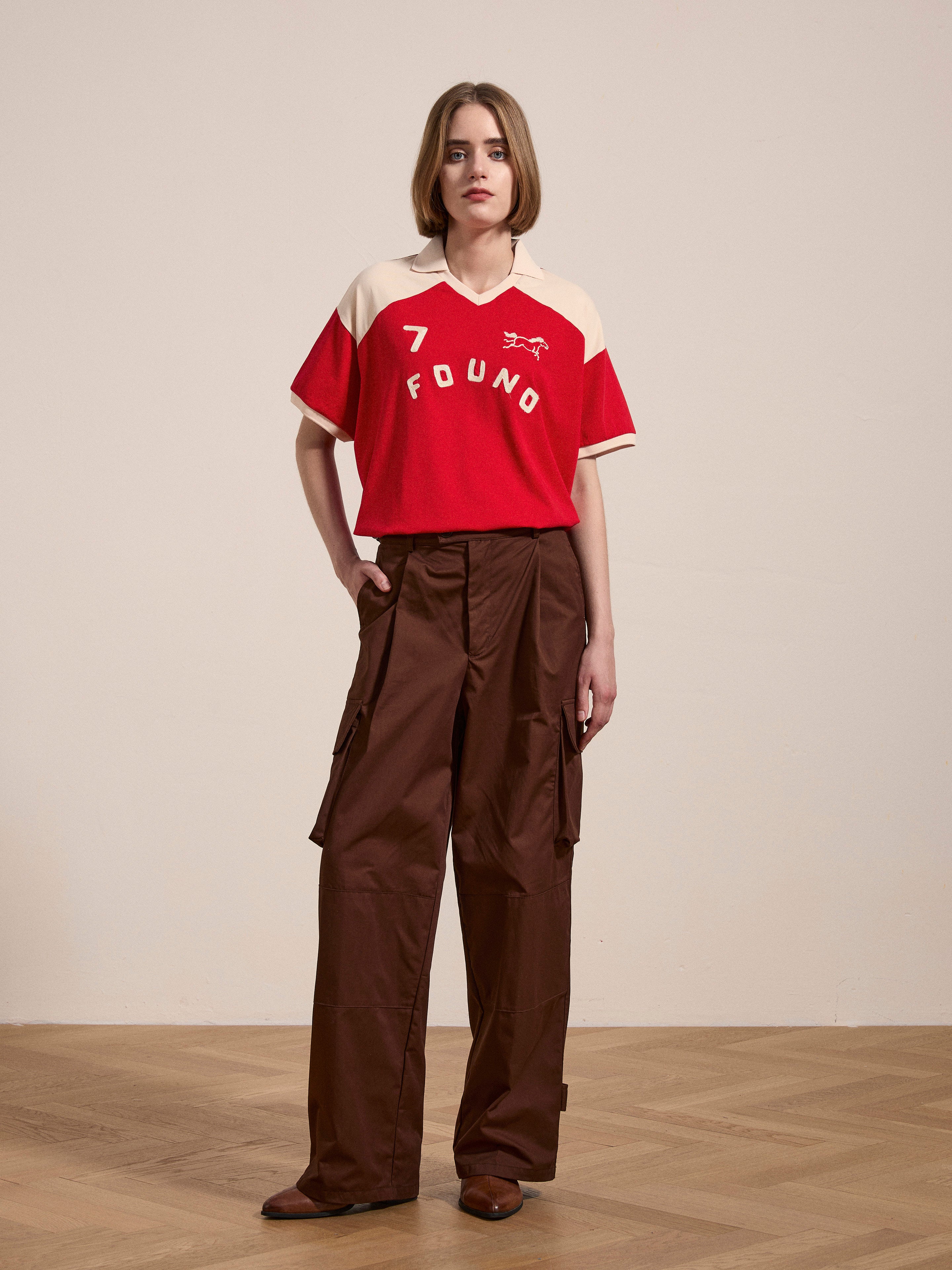 A person stands indoors wearing a Goji Red Jersey by FOUND with the text "7" and brown cargo pants, reminiscent of vintage athletic uniforms.