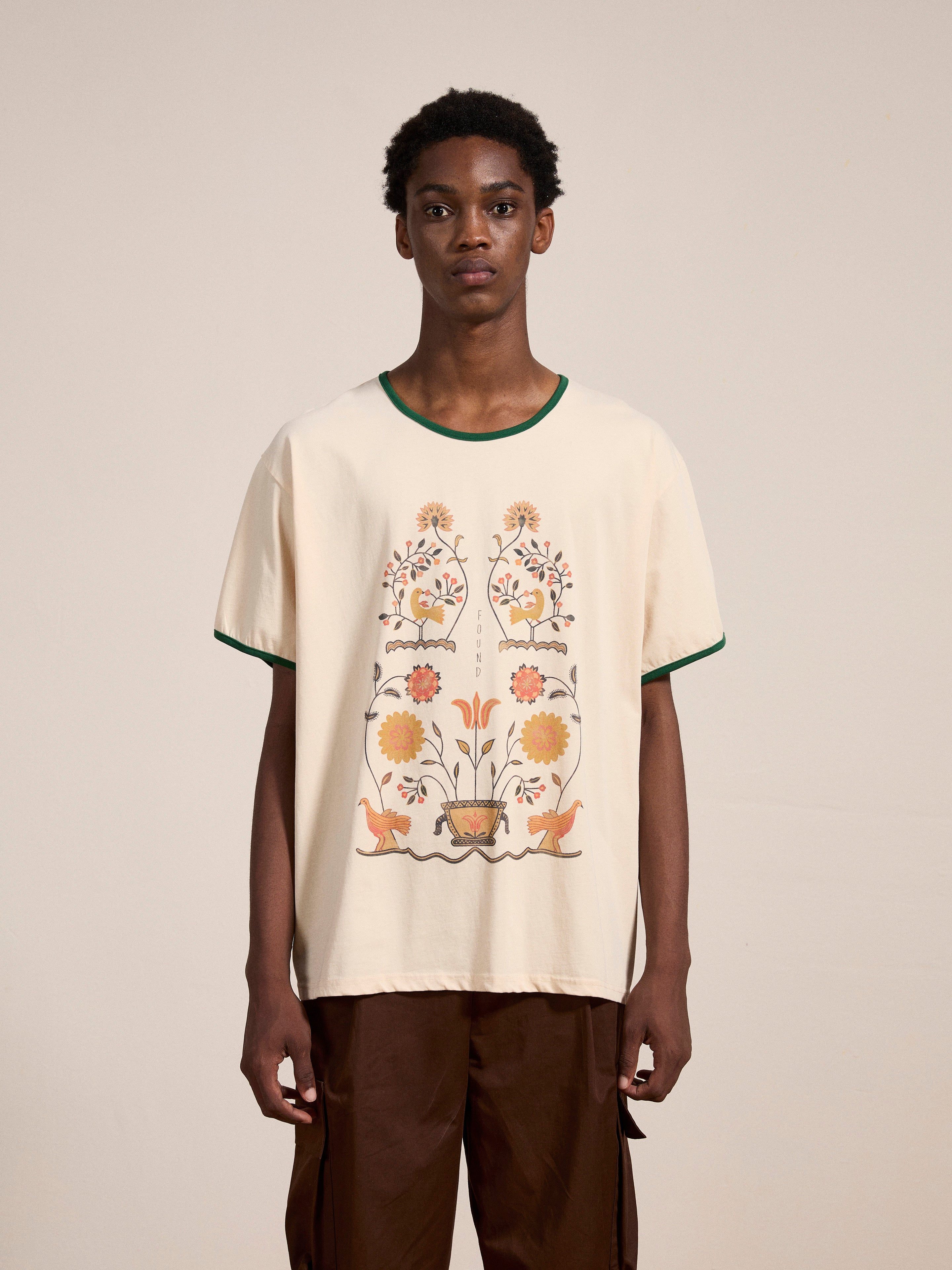 A person wearing a Cascade Floral Ringer Tee by FOUND, featuring a cream-colored base with South Asian motifs on the front, paired with brown pants, stands against a plain background.