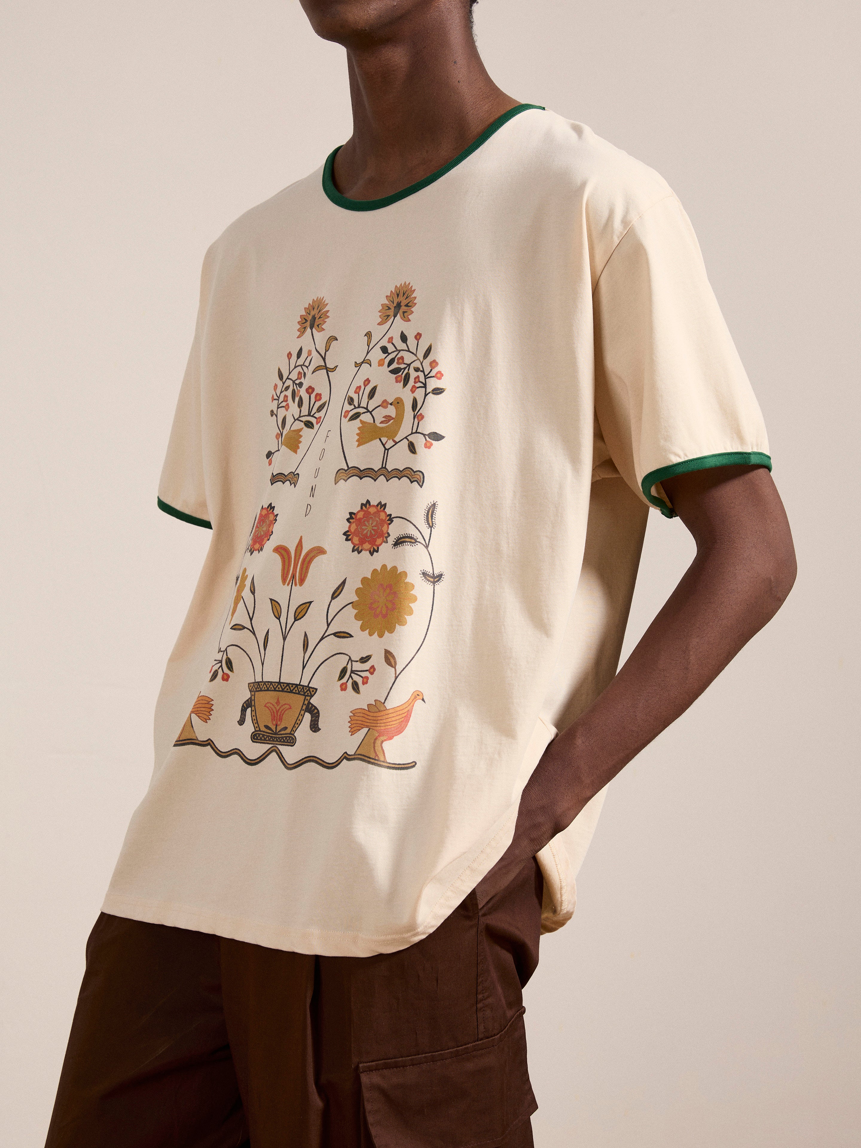 A person wearing a Cascade Floral Ringer Tee by FOUND, featuring a beige body with green trim and adorned with a floral design, paired with brown pants.