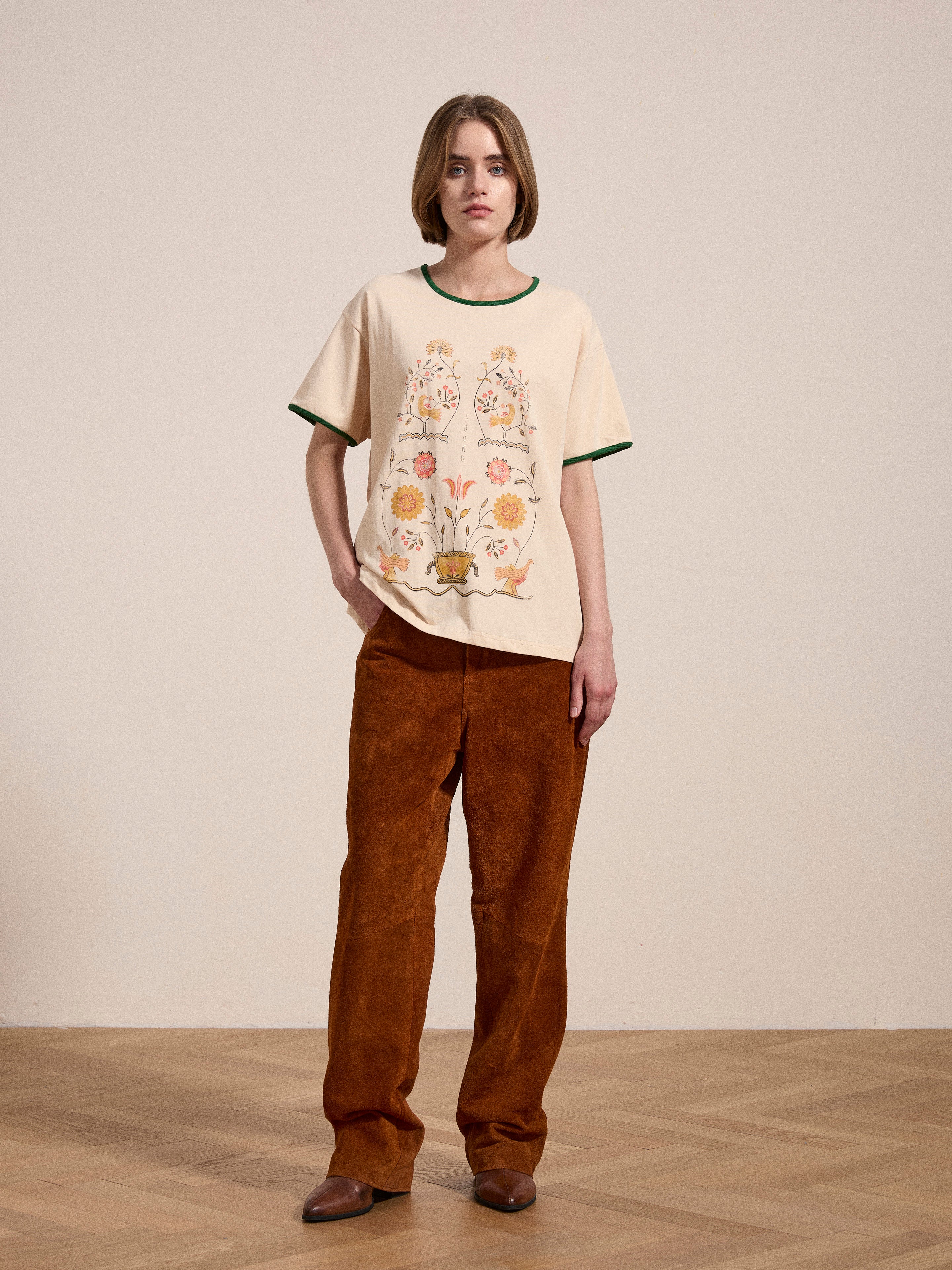 A person stands indoors on a wooden floor, wearing a Cascade Floral Ringer Tee from FOUND and brown pants. One hand is in their pocket, and they are looking forward.