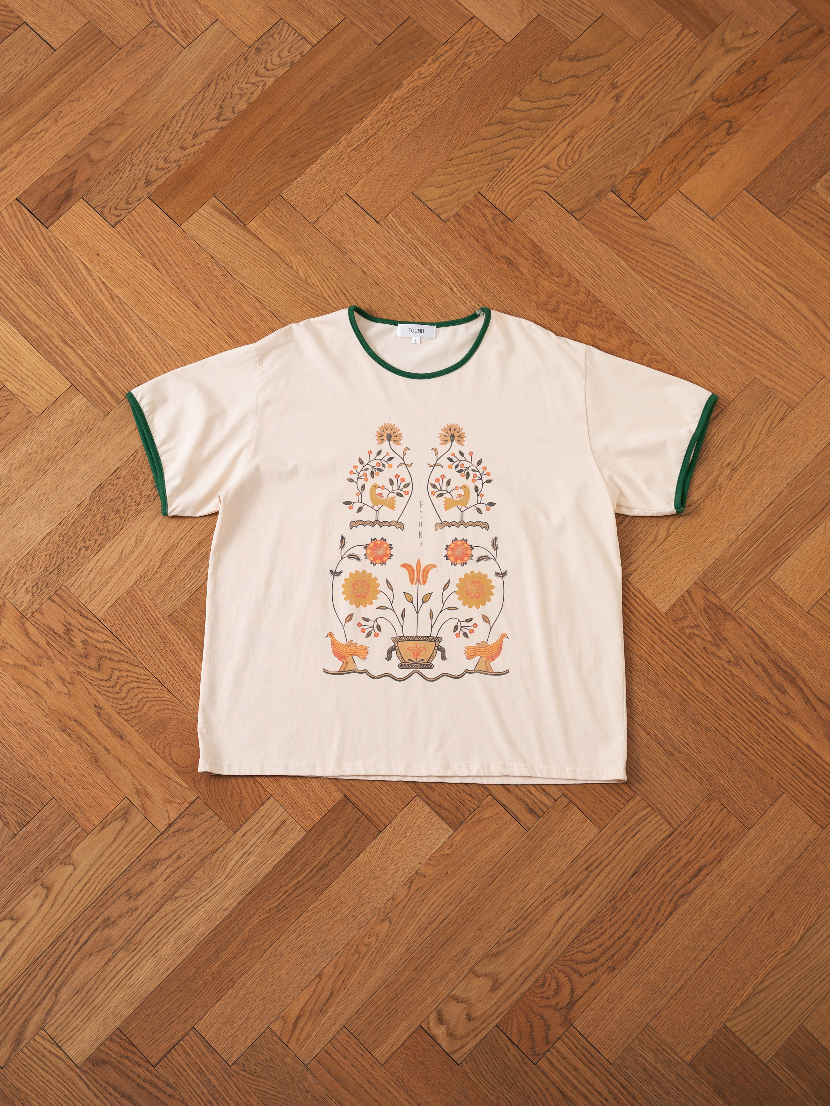 A cream-colored Cascade Floral Ringer Tee by FOUND, featuring green trim and a floral pattern inspired by South Asian motifs, laid flat on a wooden herringbone floor.