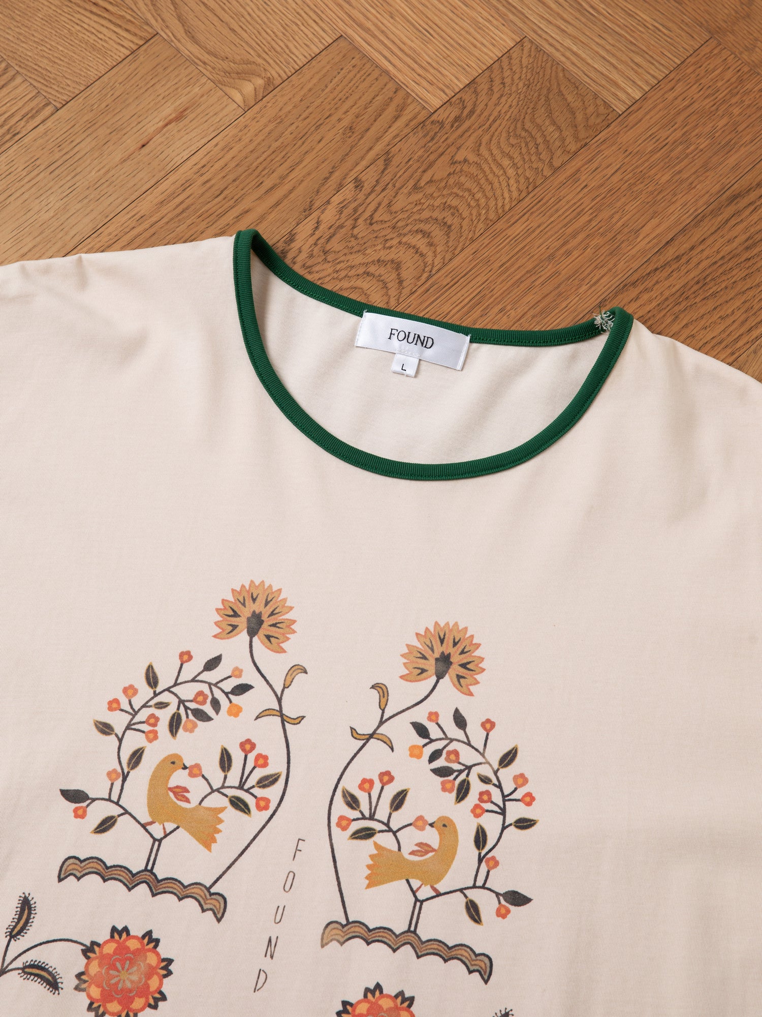 A beige Cascade Floral Ringer Tee with green trim, designed with birds, flowers, and subtle South Asian motifs, is branded with 