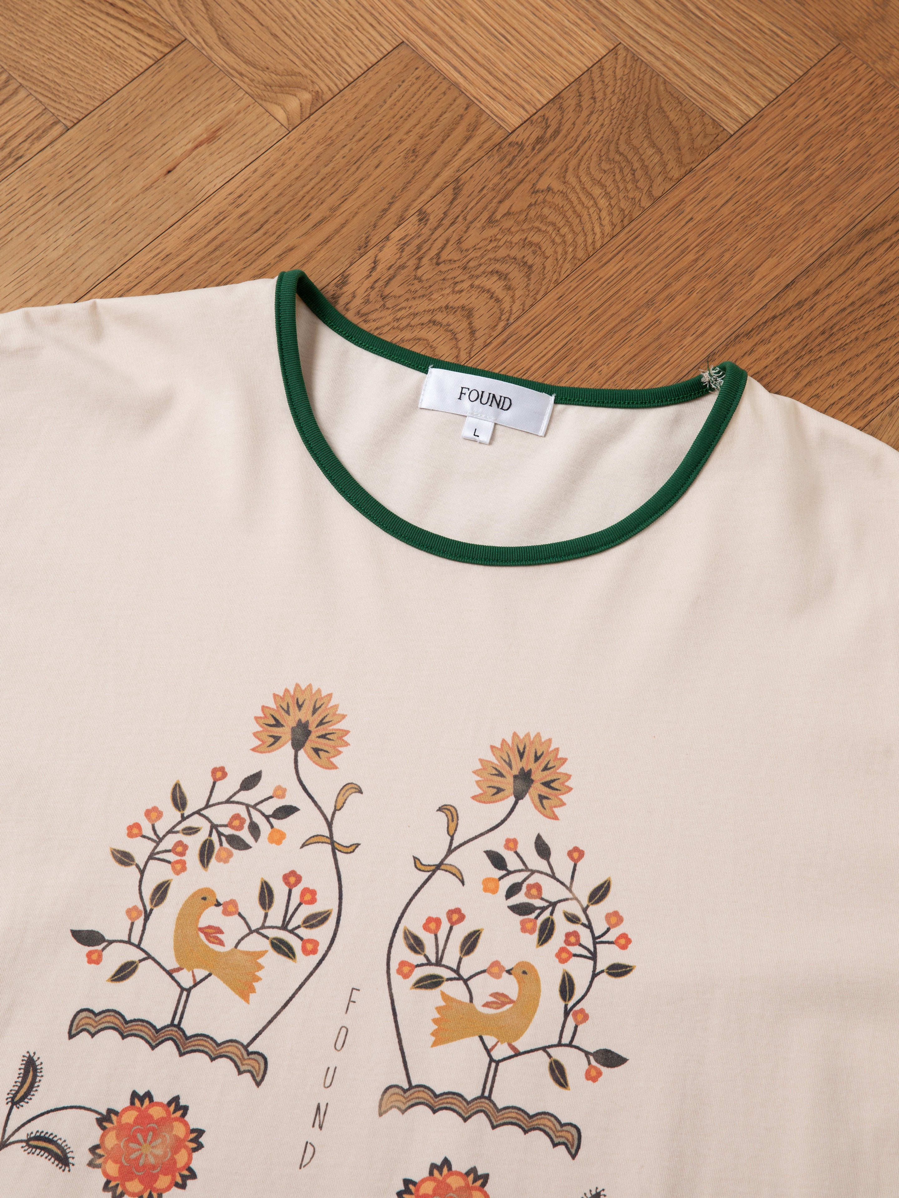 A beige Cascade Floral Ringer Tee with green trim, designed with birds, flowers, and subtle South Asian motifs, is branded with "FOUND" on the label inside the collar. The shirt is displayed on a wooden parquet floor.