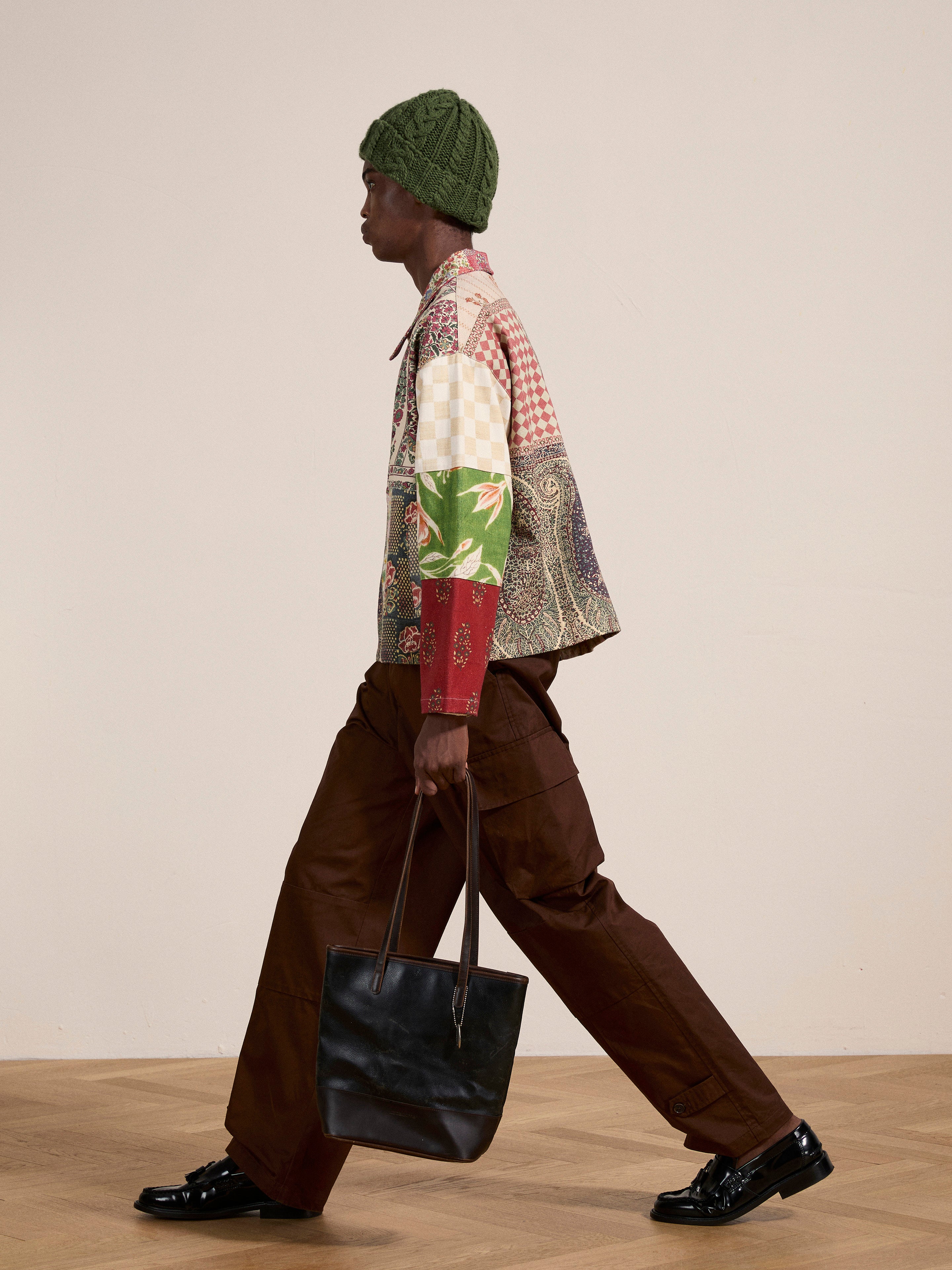 A person walking in profile, wearing a FOUND Tapestry Twill Jacket adorned with South Asian patterns, brown pants, a green knit hat, and black shoes, carrying a black tote bag in their right hand.