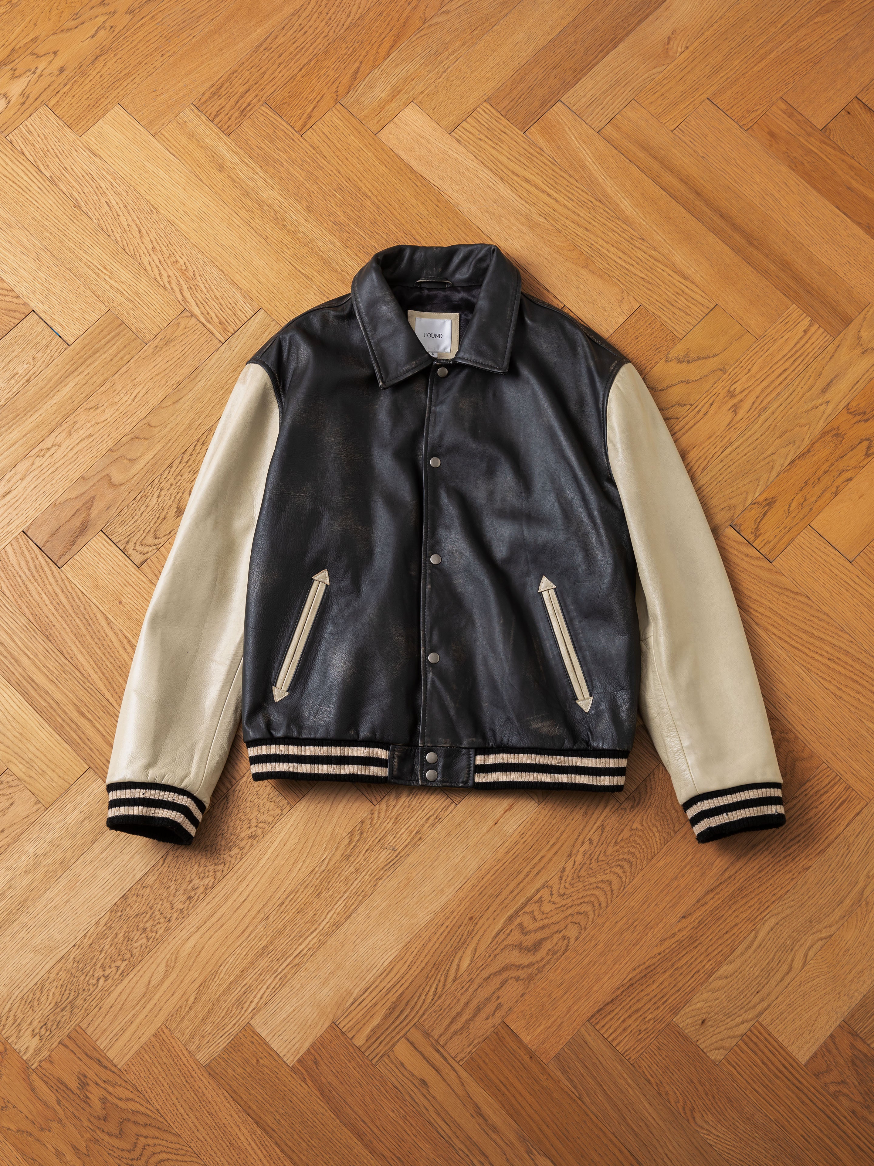 A Logan Distressed Leather Varsity Jacket by FOUND, featuring a black and white color scheme with ribbed cuffs and hem, front zipper pockets, satin lining, and a button-down closure, is laid flat on a wooden herringbone floor.