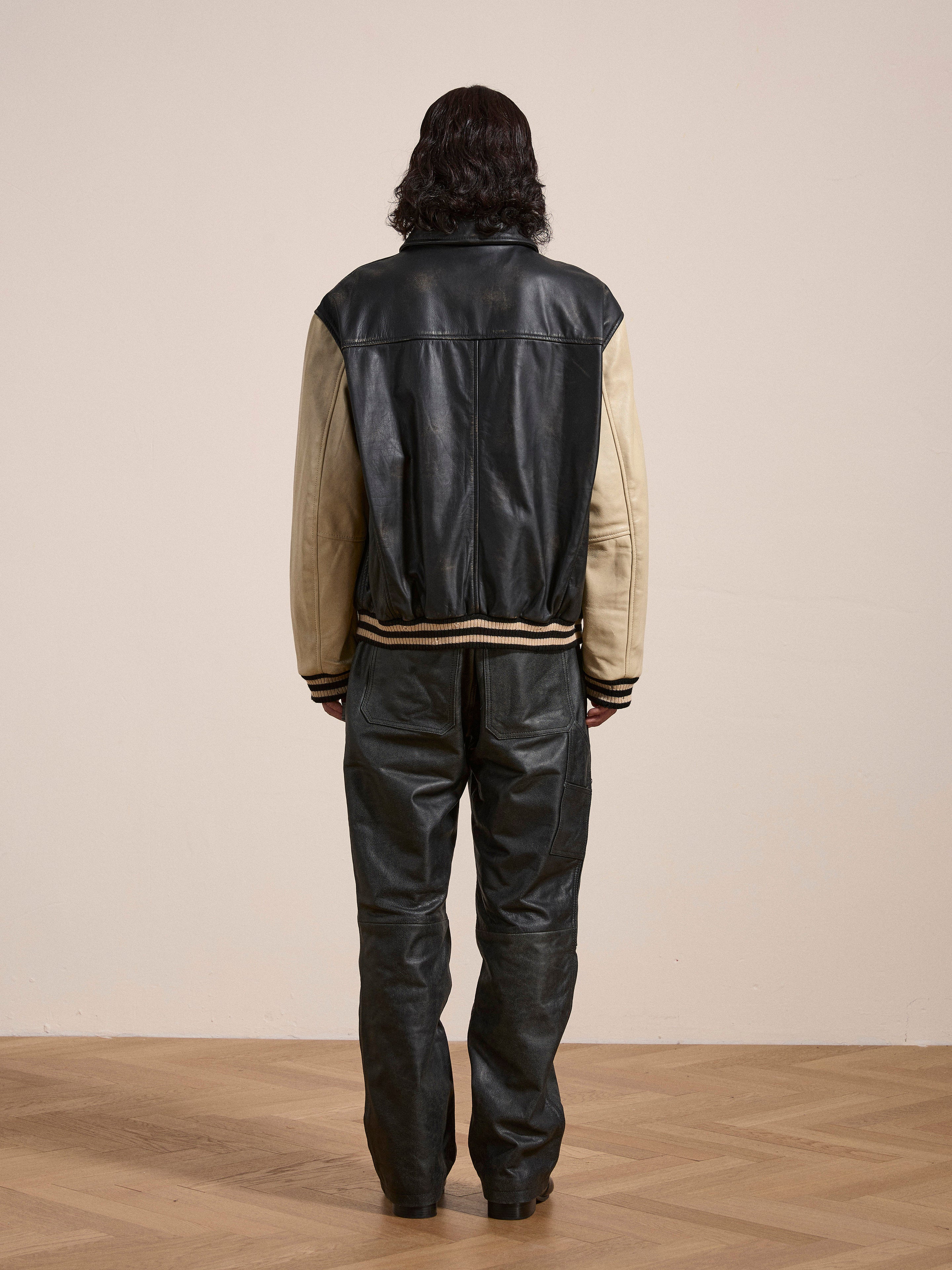 A person with long hair wearing FOUND's Logan Distressed Leather Varsity Jacket made from genuine hand-distressed leather, combined with black leather pants, stands on a wooden floor facing away from the camera.