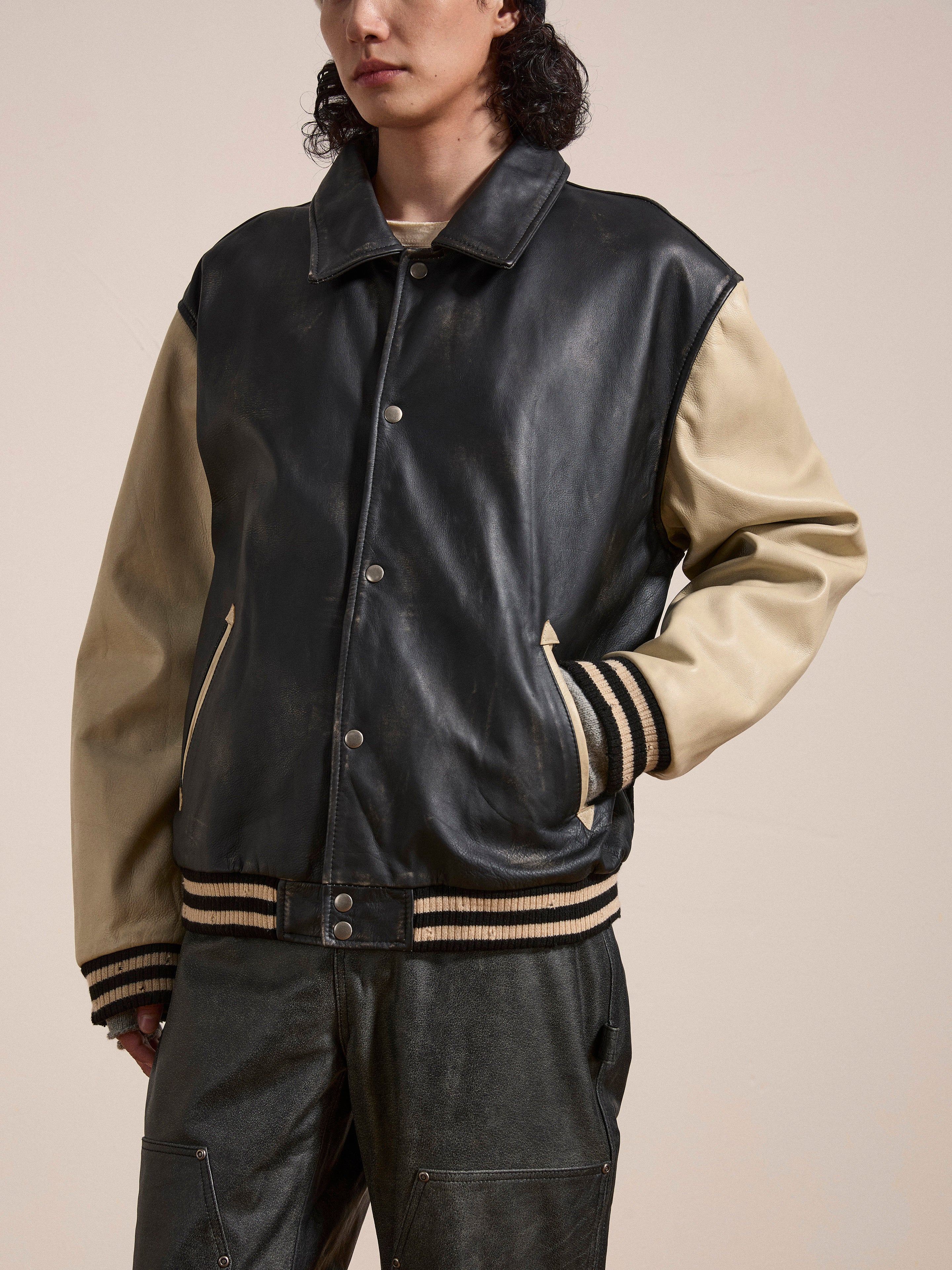 A person wearing the FOUND Logan Distressed Leather Varsity Jacket with black and beige hues, their hands in the pockets, standing against a neutral background. The genuine hand-distressed leather and luxurious satin lining make this piece truly standout.