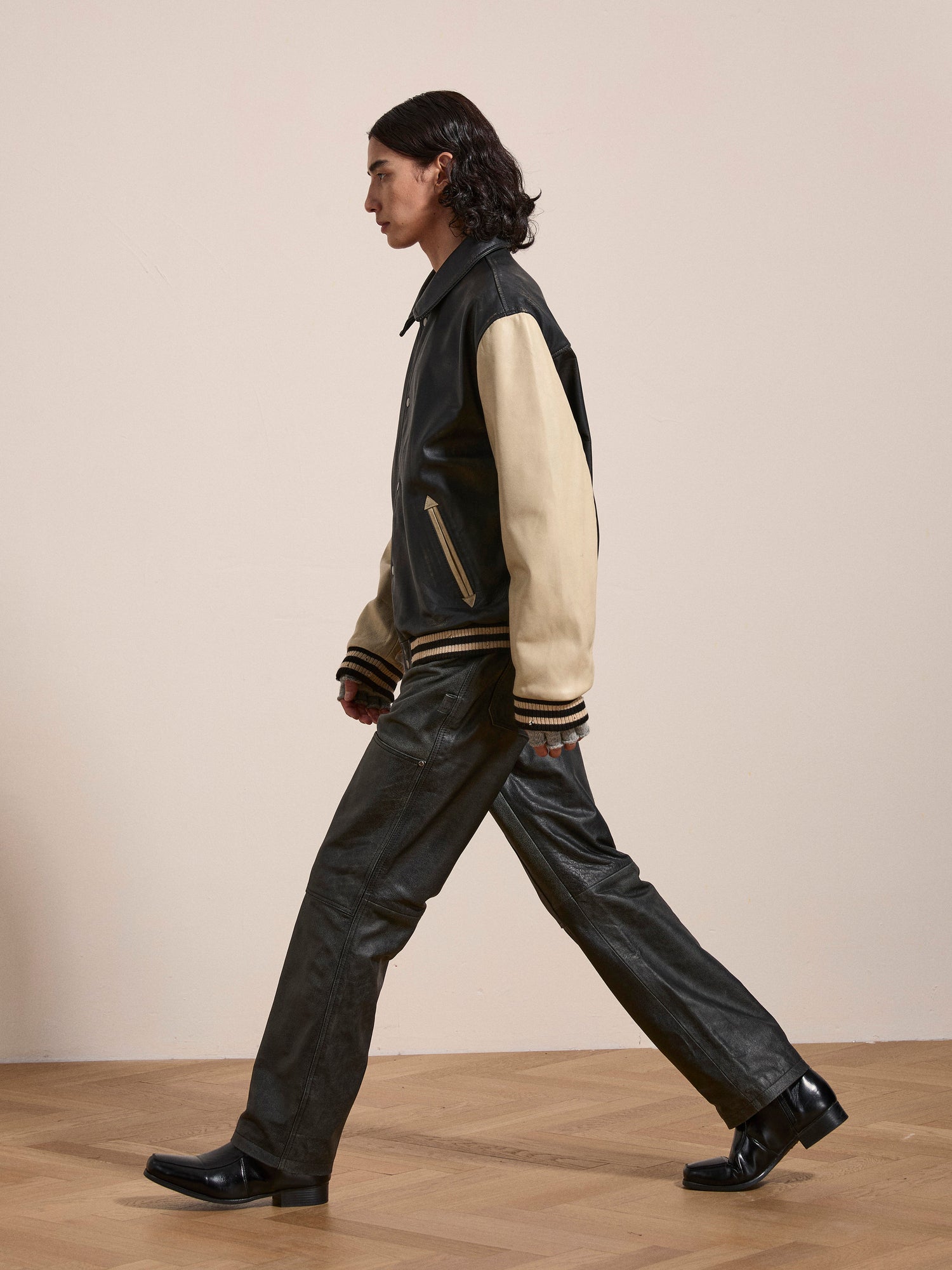 A person with long dark hair walks indoors wearing the FOUND Logan Distressed Leather Varsity Jacket, leather pants, and black shoes. The jacket, made of genuine hand-distressed leather with a satin lining, complements the edgy profile view.